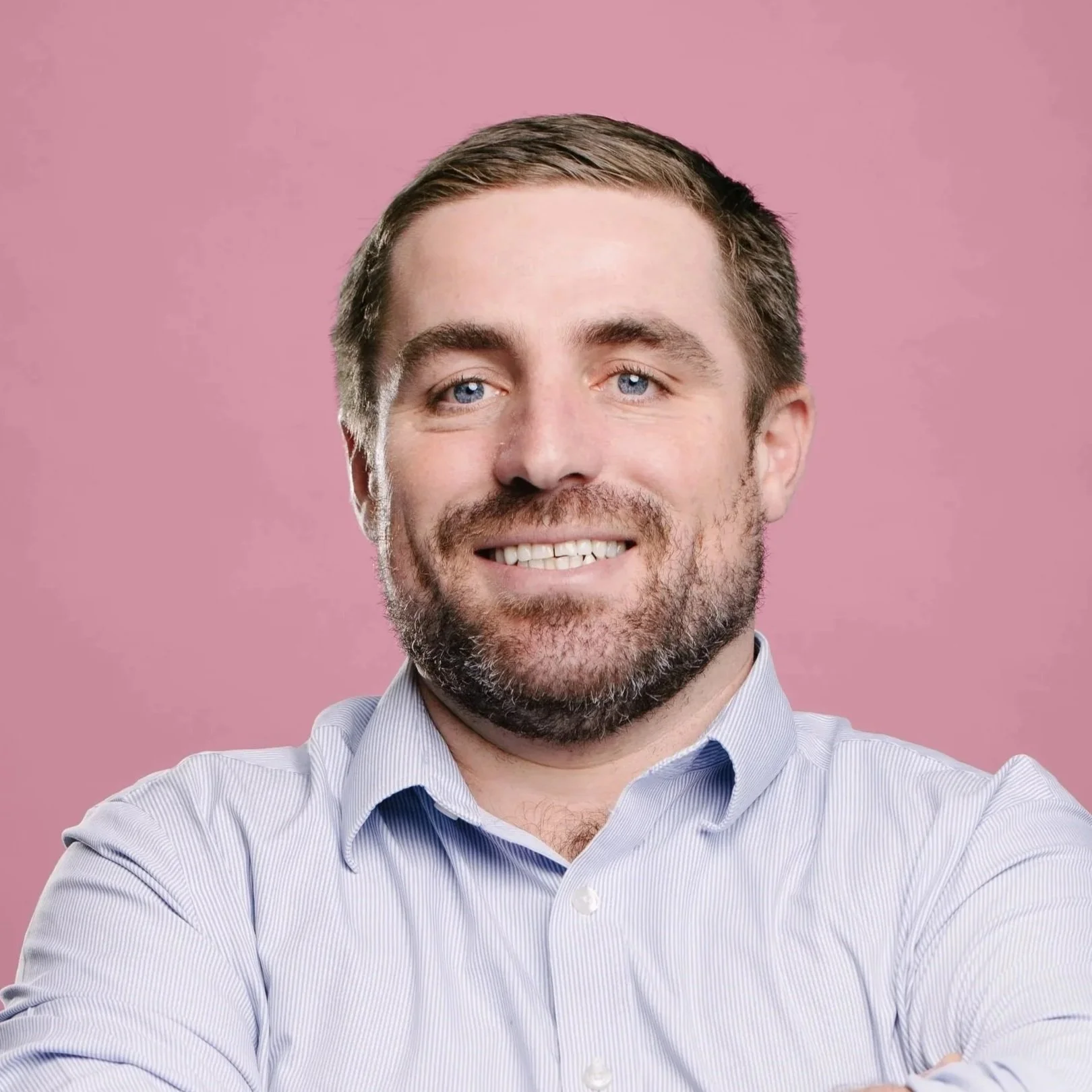 A white man with brunette hair and a light beard and mustache smiles at the camera. He wears a white collared shirt and dark blue tie with a tan wall and window in the background.