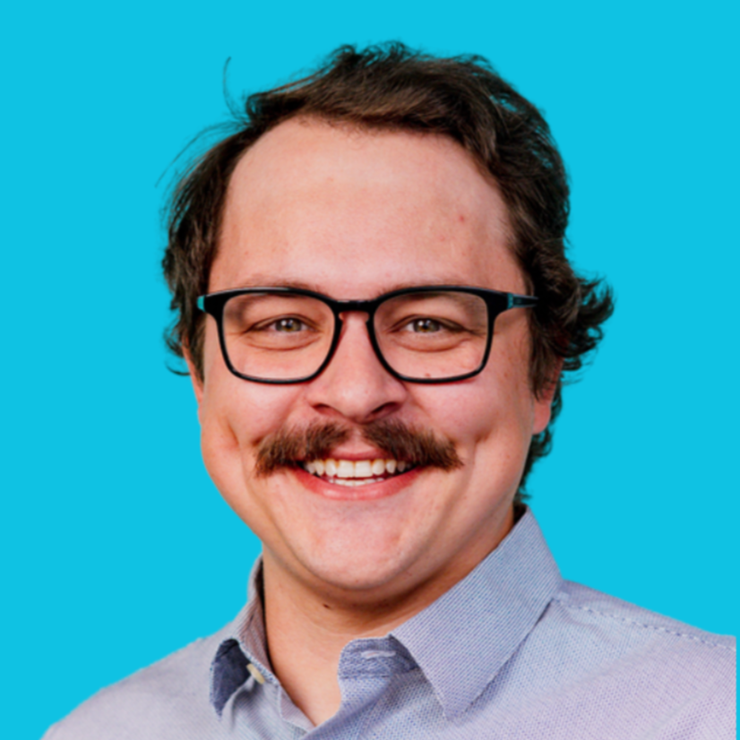 A white man in a blue collared shirt smiles at the camera. He has short brown hair, black glasses, and a mustache.