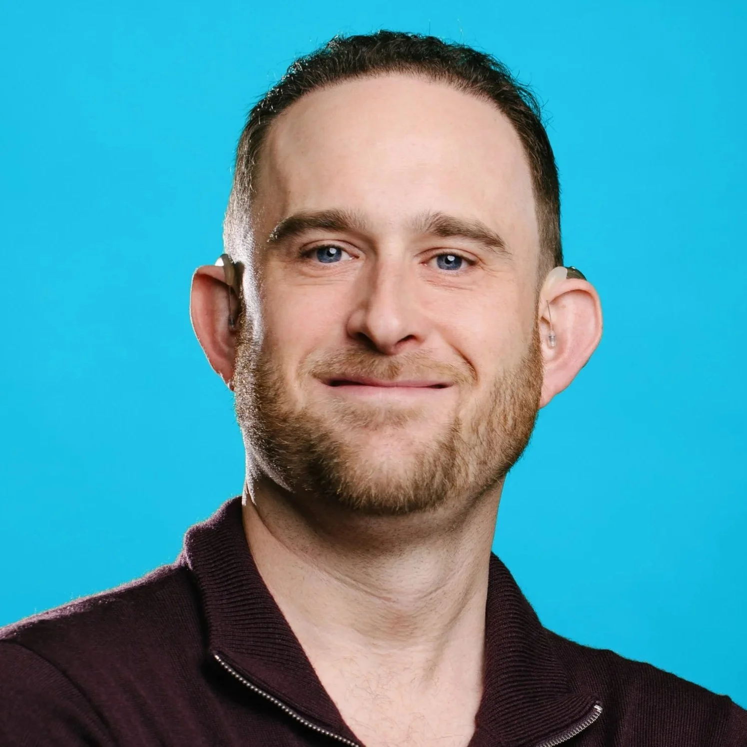 A white man with short brown hair and a trimmed beard, smiling in a navy blazer against a vertical striped wall.