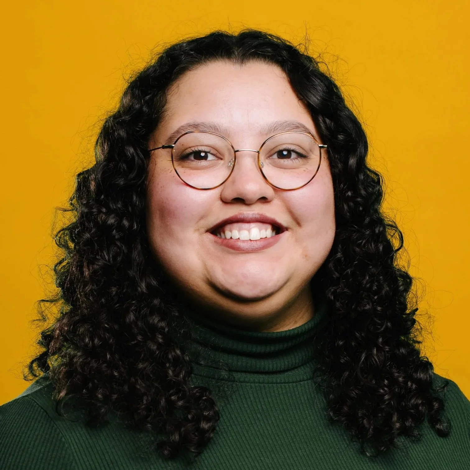 A Black woman with medium-toned skin and long curly hair, smiling softly in a navy shirt, in front of a dark grey background.