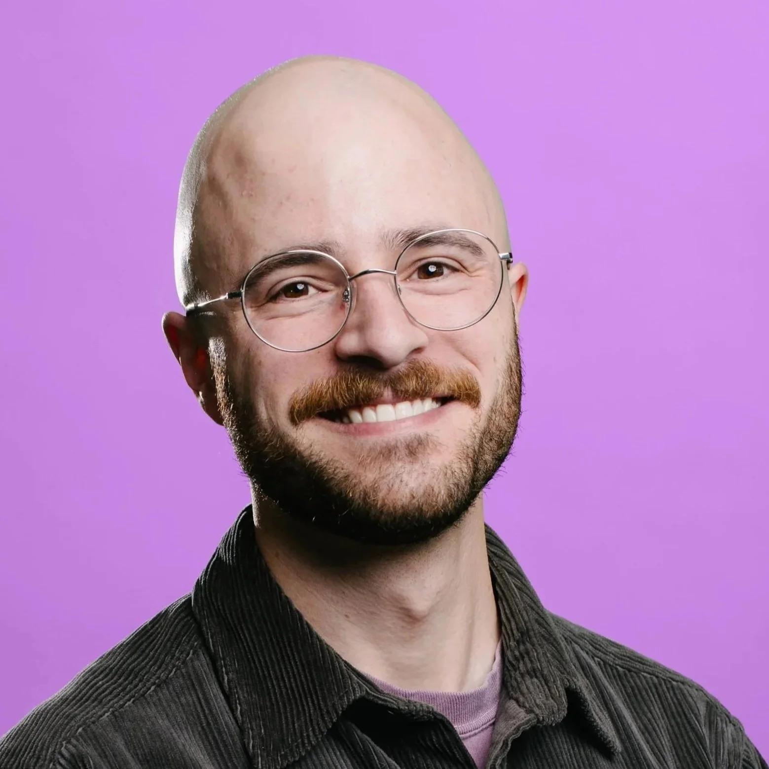 A bald white man with a dark brown beard and mustache wearing clear glasses smiles at the camera. He wears a striped shirt and collared dark blue jacket with a nature background.