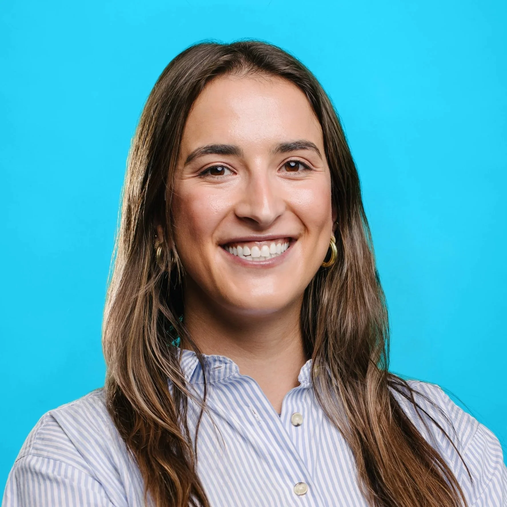 A brunette white woman smiles at the camera with a nature background behind her.