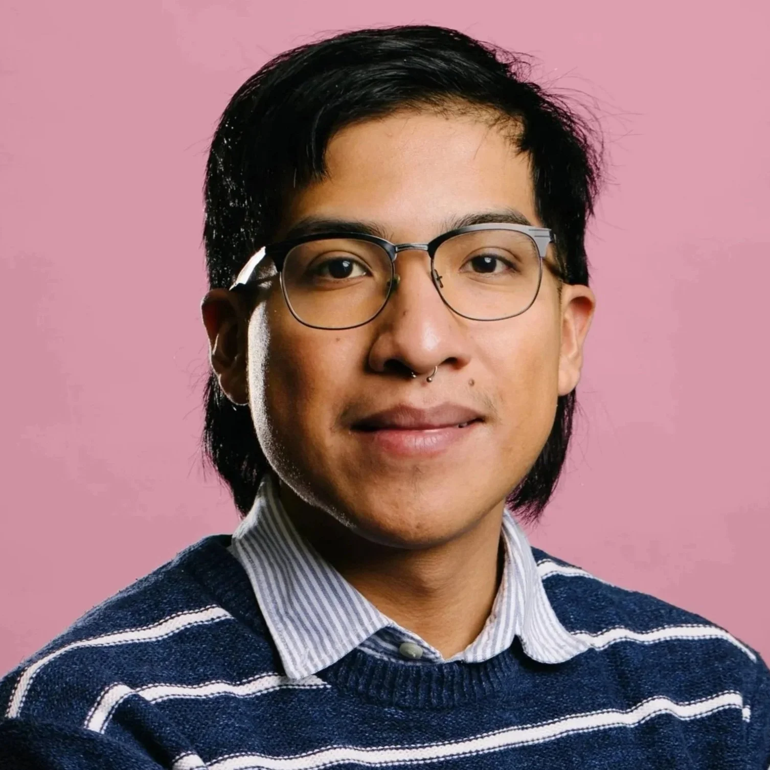 A Latino man looks at the camera/ He has medium length dark hair, glasses, and wears a dark blue and thin white striped shirt with a light blue collar