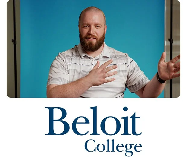 A white man with light brown short hair, beard, and mustache wearing a suit and white and blue checkered shirt close-mouthed smiles at the camera in front of a yellow background with the Beloit College logo below him.