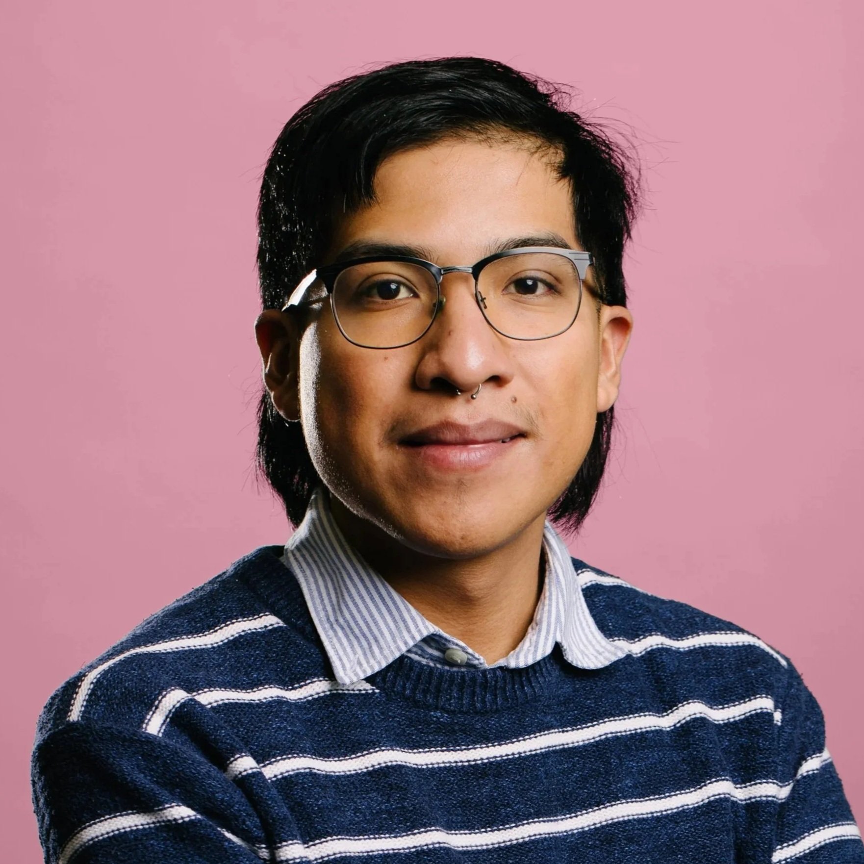 A Latino man looks at the camera/ He has medium length dark hair, glasses, and wears a dark blue and thin white striped shirt with a light blue collar