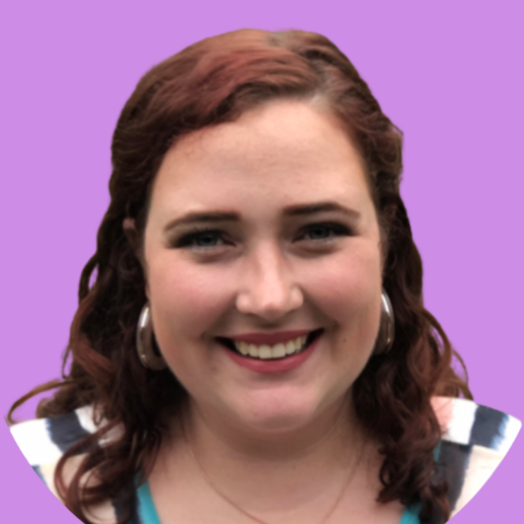 A white woman with wavy brunette hair smiles directly at the camera. She wears small hoop earrings and a necklace.