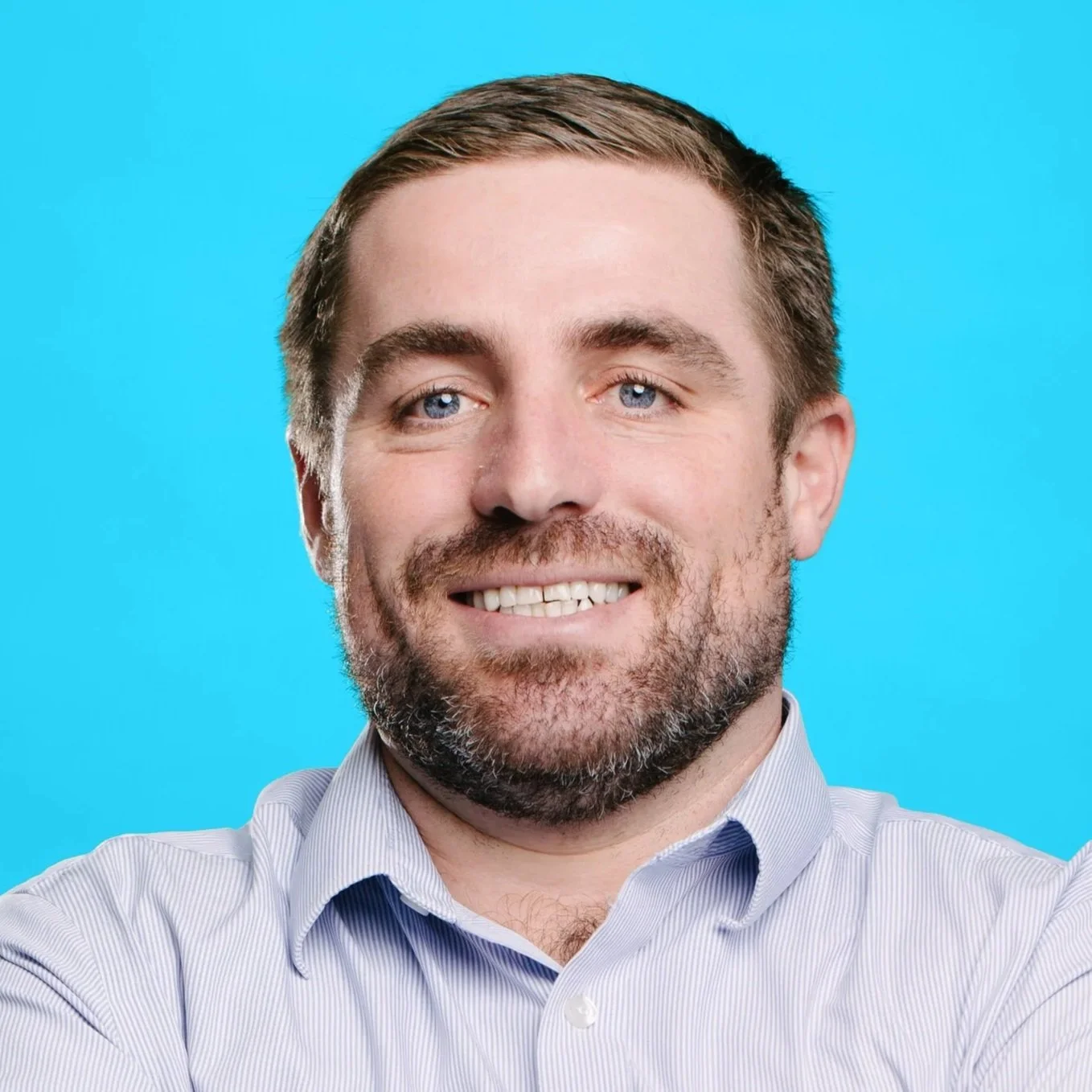 A white man with brunette hair and a light beard and mustache smiles at the camera. He wears a white collared shirt and dark blue tie with a tan wall and window in the background.