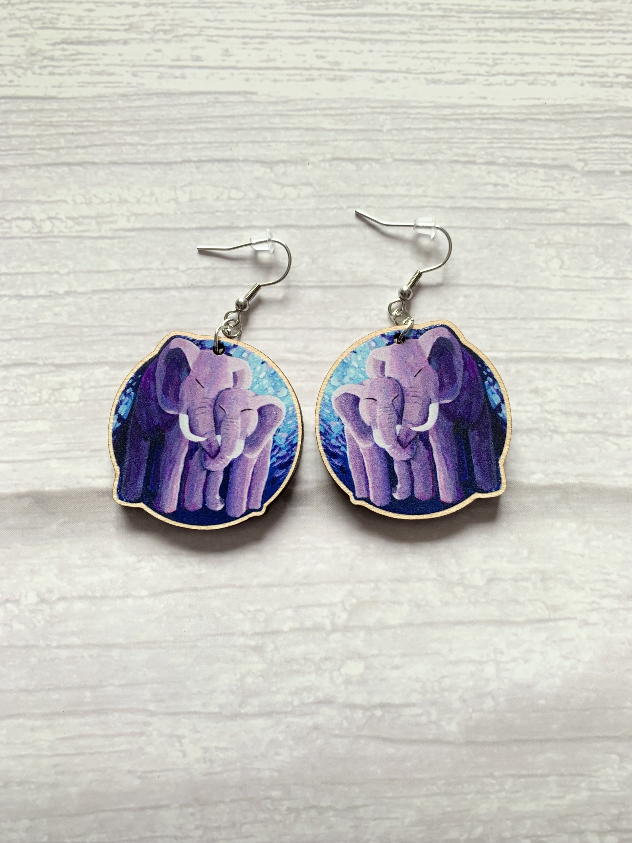 Two hook back, drop style earrings are laying on a wooden background. The earring design is of die cut elephants cuddling.