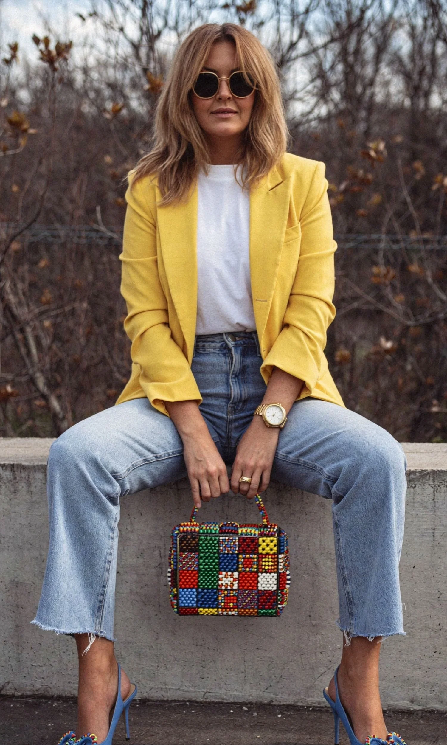 Woman sitting on a concrete ledge outdoors, wearing a yellow blazer, white t-shirt, jeans, and blue high heels, holding a colorful beaded bag, with sunglasses and a gold watch, in front of leafless bushes.