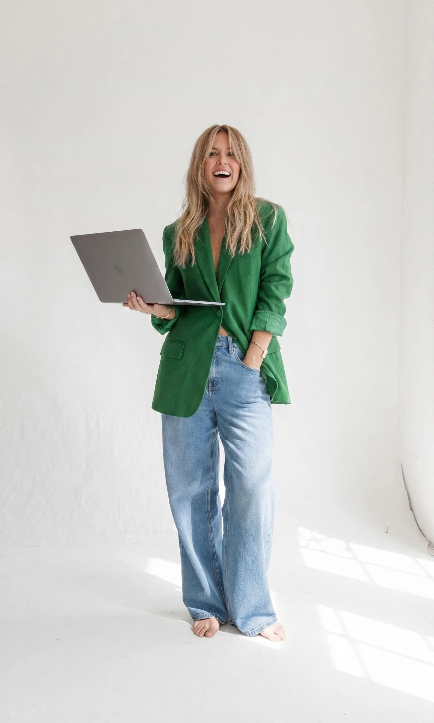 A woman with long blonde hair wearing a green blazer and baggy jeans is smiling and holding a laptop while standing barefoot on a white background.