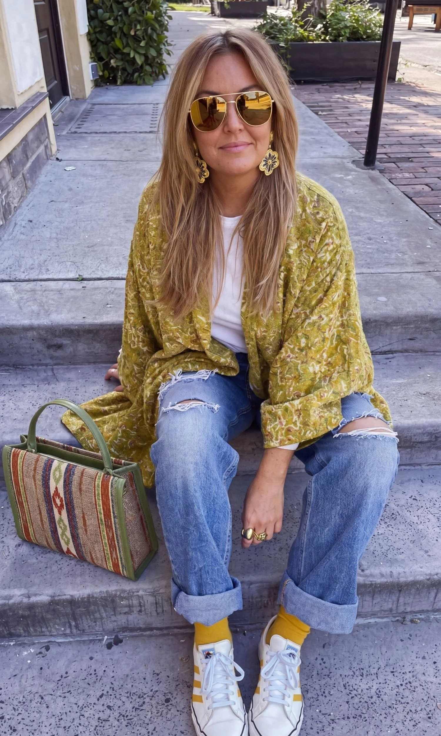 A woman with long blond hair wearing sunglasses, yellow floral earrings, a yellow textured jacket, a white t-shirt, ripped blue jeans, yellow socks, and white sneakers with yellow stripes, sitting on steps outside with a striped bag beside her.