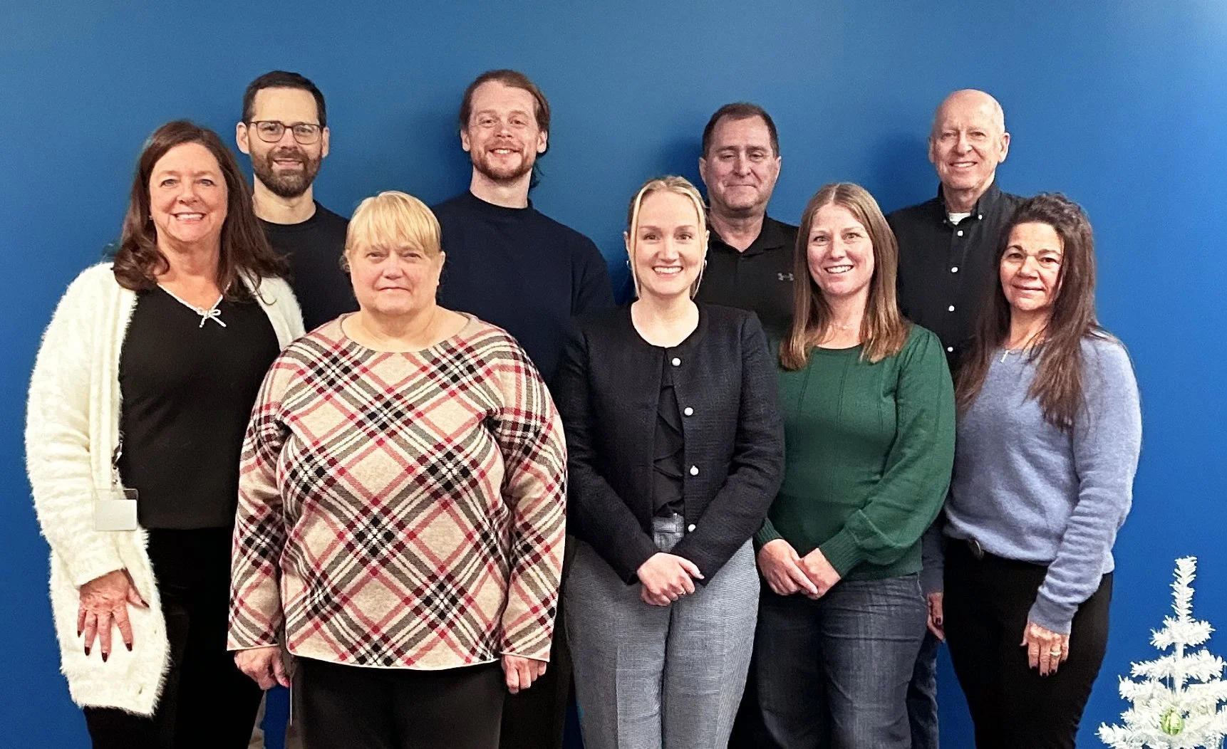Staff photo of IFPTI; nine employees in front of a blue wall with a tiny Christmas tree to the side