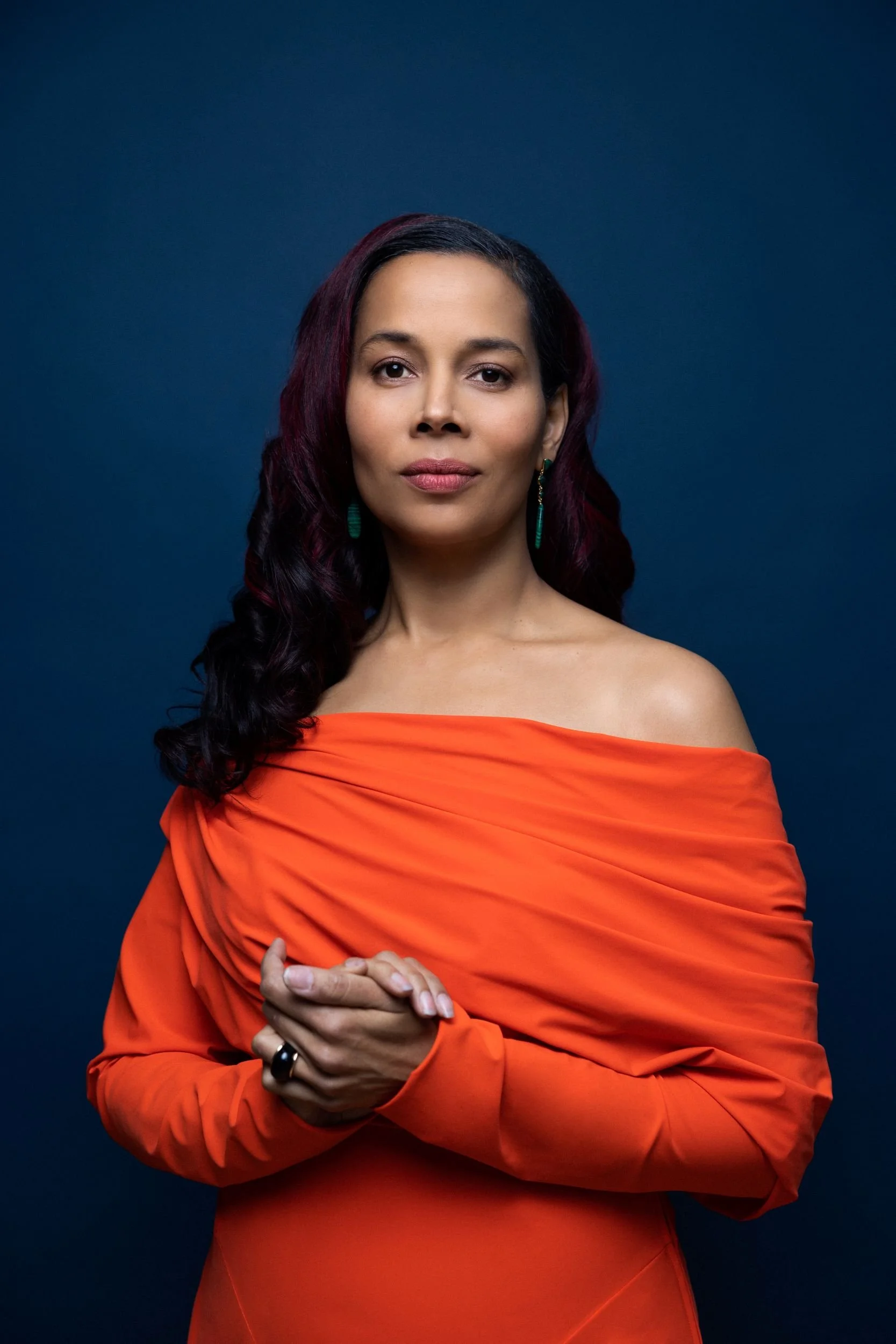 A woman with dark hair and wearing teal earrings is dressed in an off-the-shoulder orange gown, standing against a dark blue background.