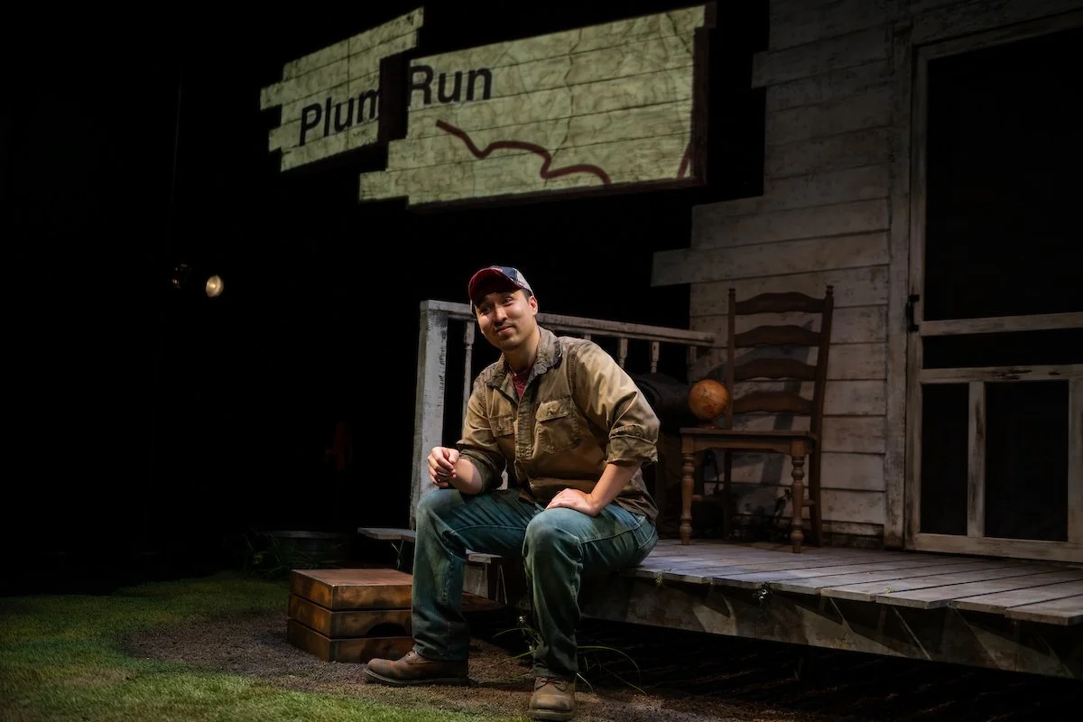 A man in casual work clothes, including a tan jacket and a baseball cap, is sitting on a small wooden stool on a stage set designed to look like an outdoor porch or rustic cabin. Behind him is a wooden chair and a globe, with a large sign that reads "Plum Run" hanging above. The setting has a weathered wood backdrop and looks like a theatrical or stage production.