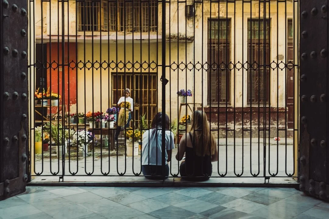 The iron gates of a church in Havana open directly onto the street, where life moves at its usual pace. Just inside, two women sit quietly on the steps, their backs to the camera. Beyond them, a flower vendor carefully arranges sunflowers and other b