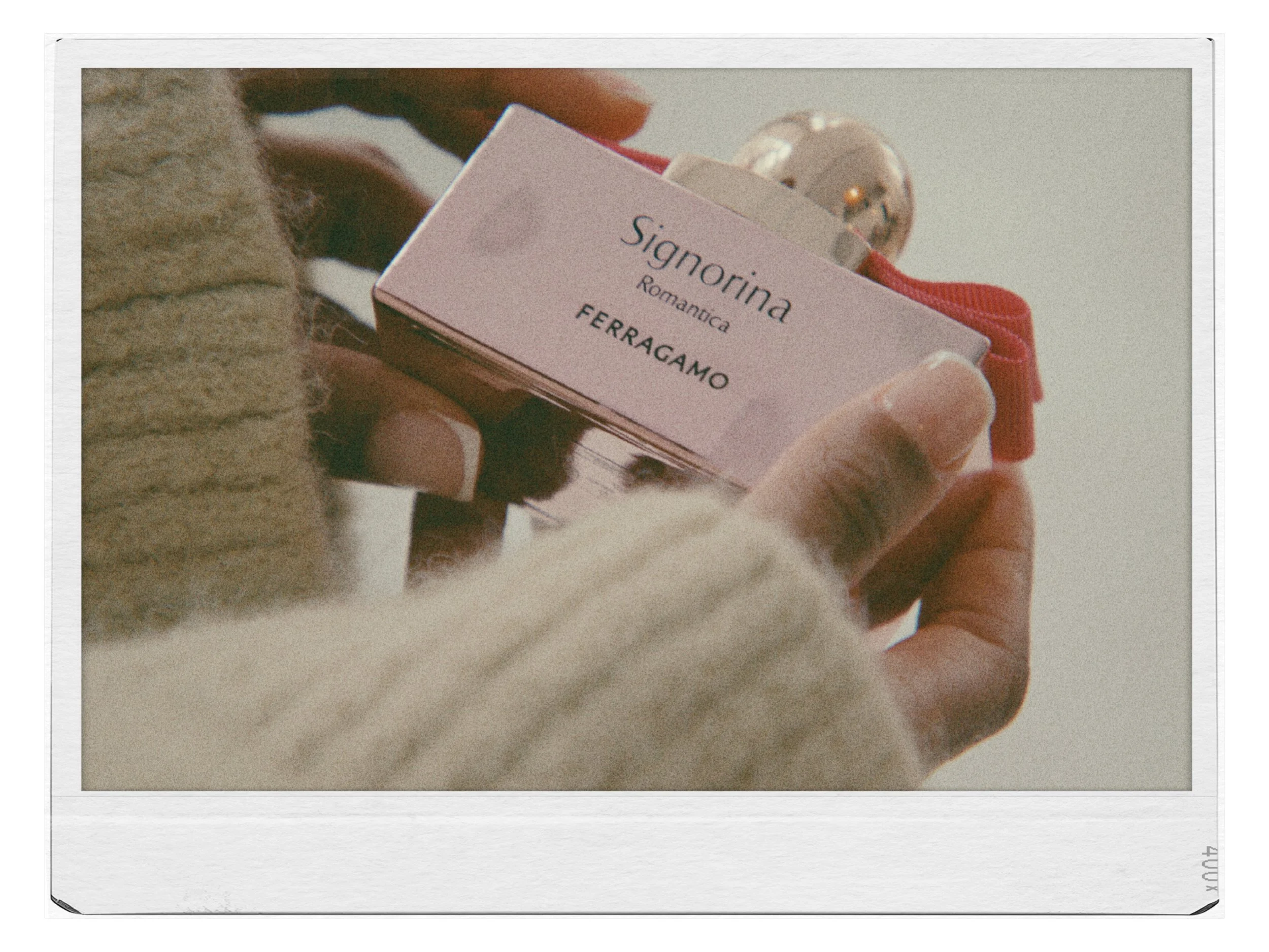 Close-up of a person holding a small pink box of Signorina Romanica perfume by Ferragamo, with a red ribbon and a shiny spherical decoration attached.