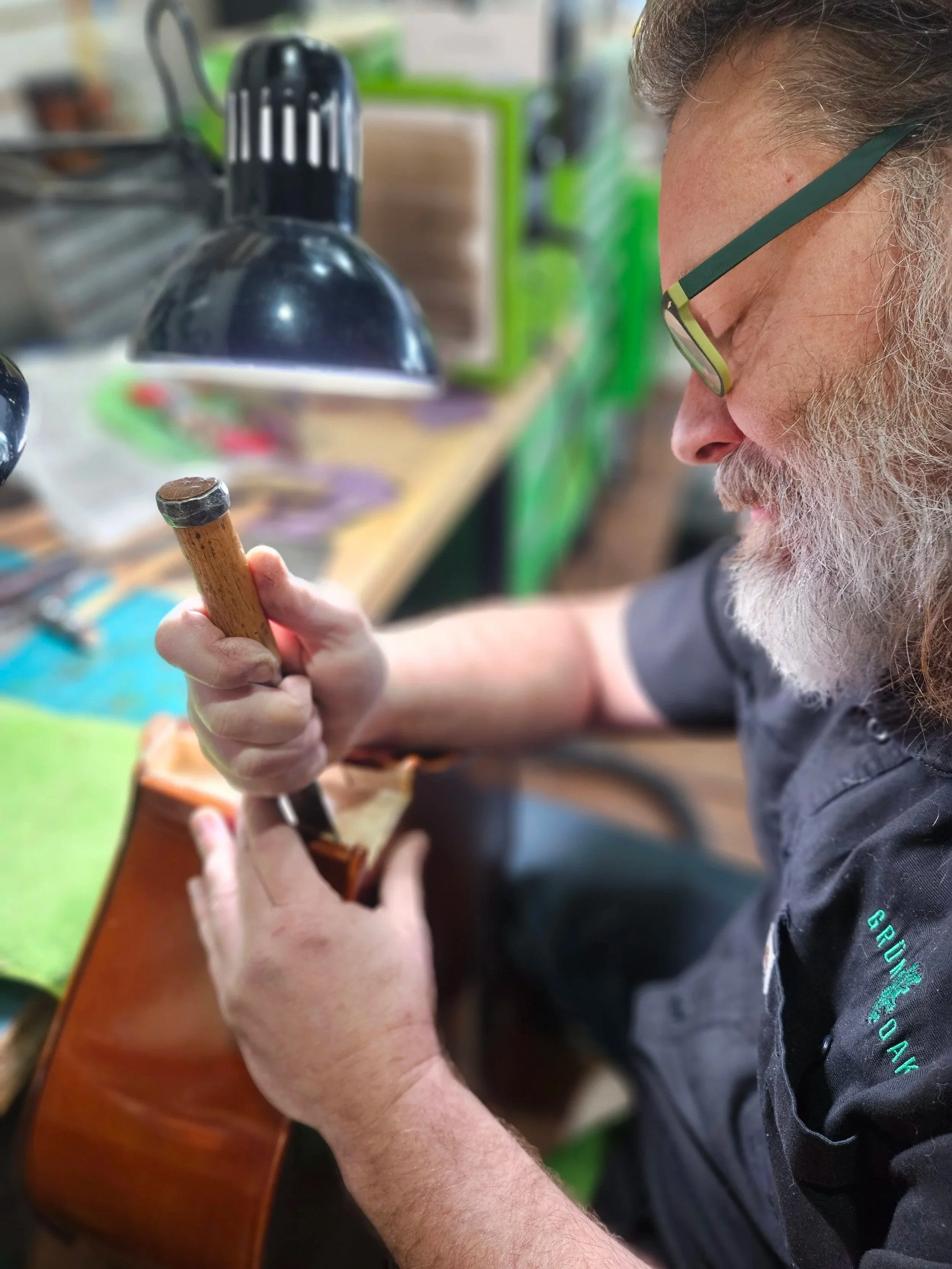 A man with glasses and a gray beard is crafting a guitar in a workshop, using a hammer.