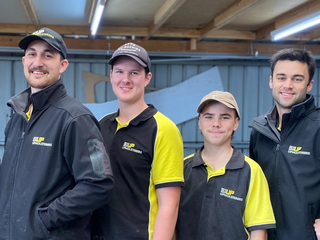 Four men standing together inside a workshop, all wearing black and yellow uniforms with 'EyreUP Upholsterers' logos, smiling at the camera.