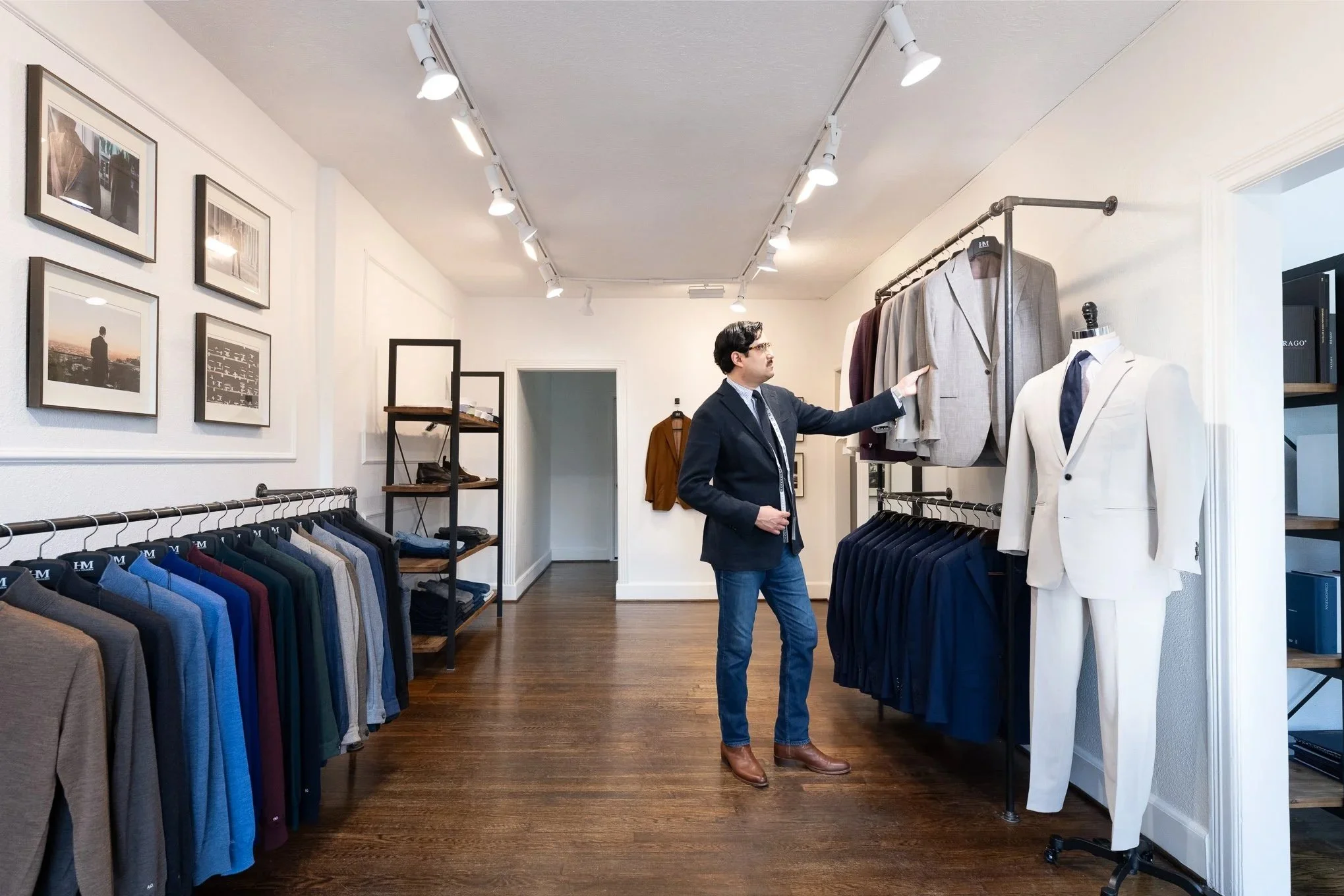 A man in a dark blazer, jeans, and brown shoes is browsing men’s suits in a clothing store. He is standing next to racks of suits and jackets, with a white suit on a mannequin to his right.