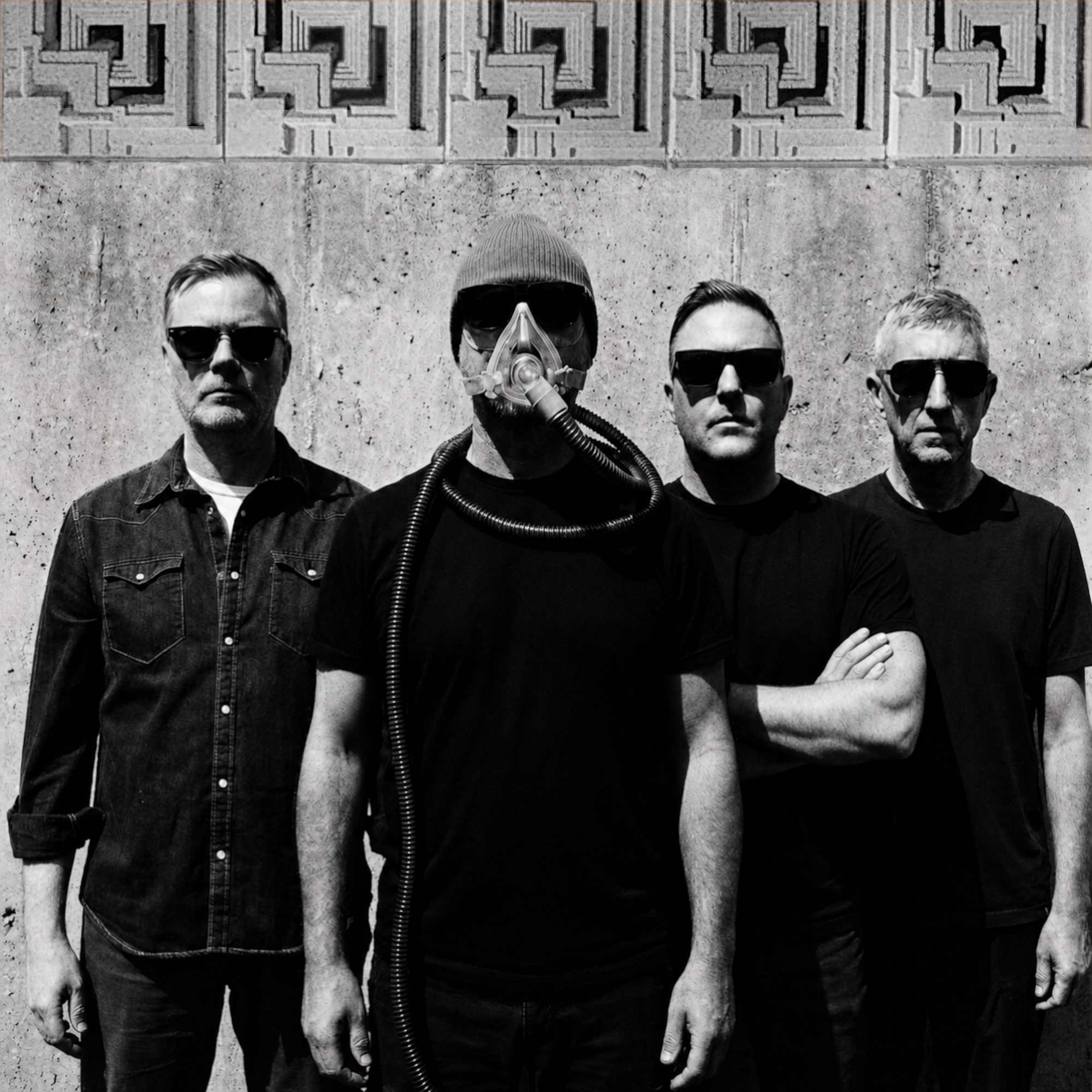 Four men standing side by side outdoors against a textured wall, one wearing a mask and breathing apparatus, the others wearing sunglasses, all dressed in dark clothing.