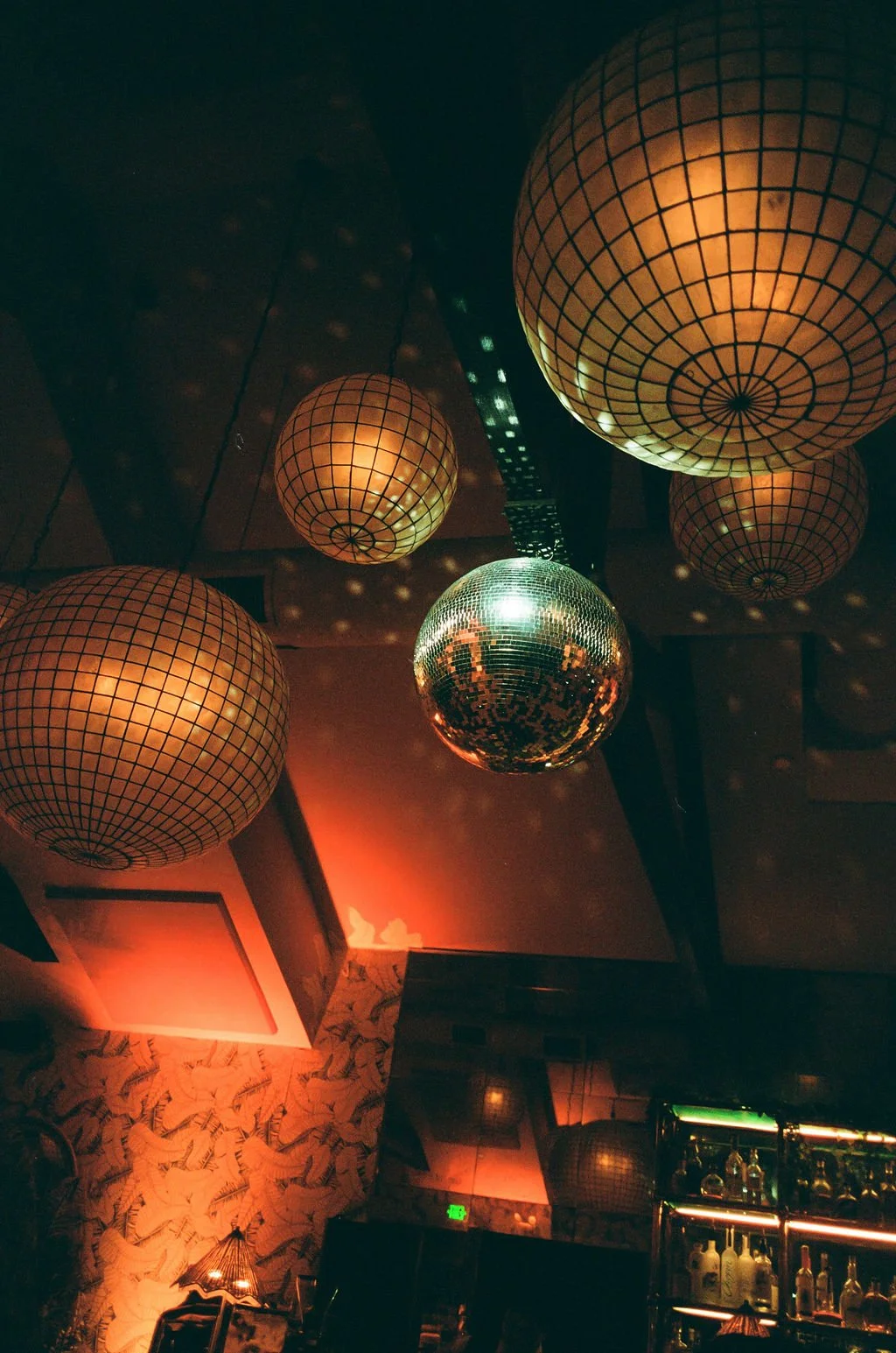 Lighting fixtures including paper lanterns and a disco ball hanging from a ceiling inside a dimly lit room, with patterned wallpaper and a bar area in the background.