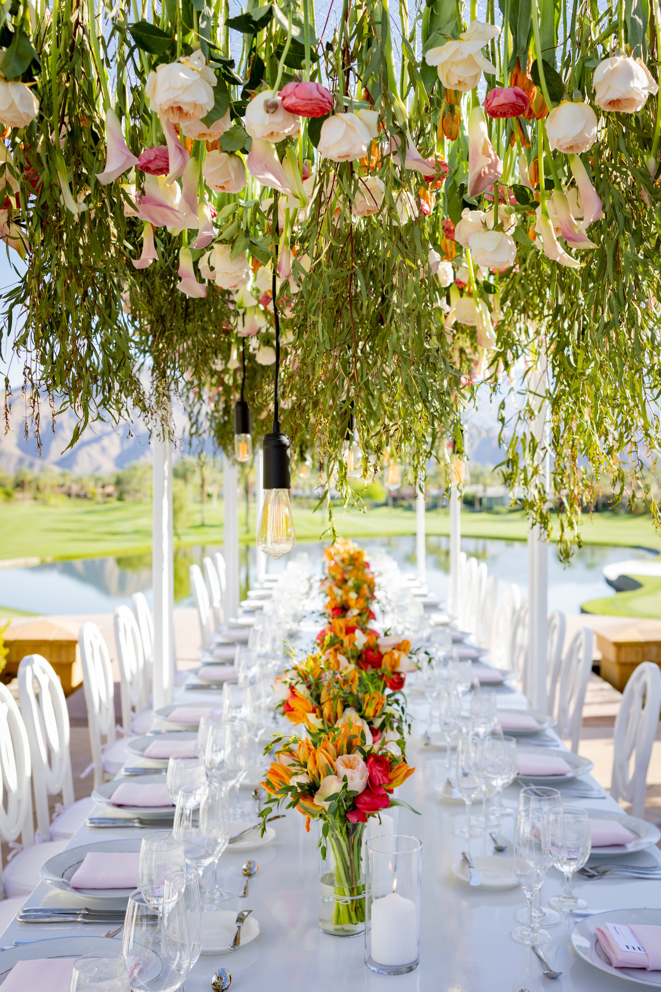 Outdoor wedding reception with a long table decorated with a colorful floral centerpiece, white chairs, and hanging floral arrangements above, set against a scenic backdrop of a pond, green grass, and mountains.