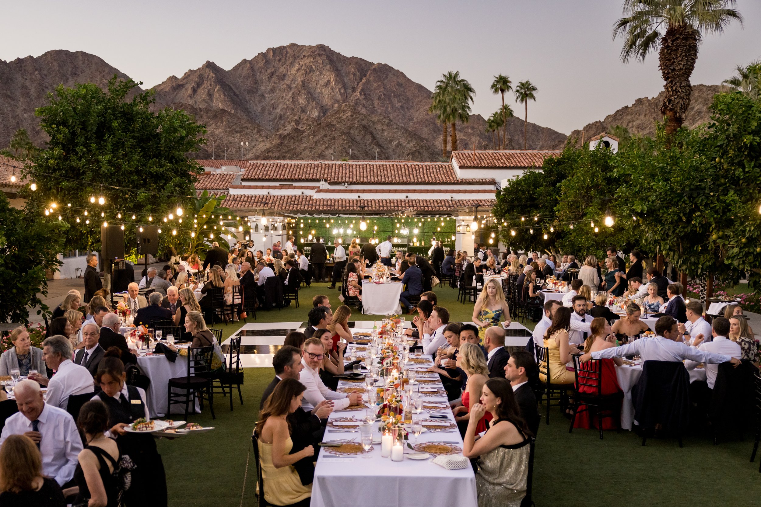 Outdoor banquet with many people seated at tables, string lights overhead, mountains in the background, and trees and palm trees around the event area as the sky transitions from day to evening.