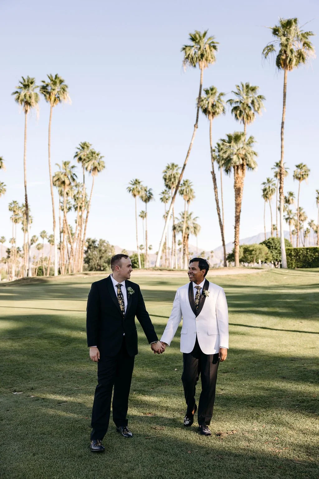 Two men in wedding attire holding hands and walking on a grassy area with tall palm trees and mountains in the background on a sunny day.