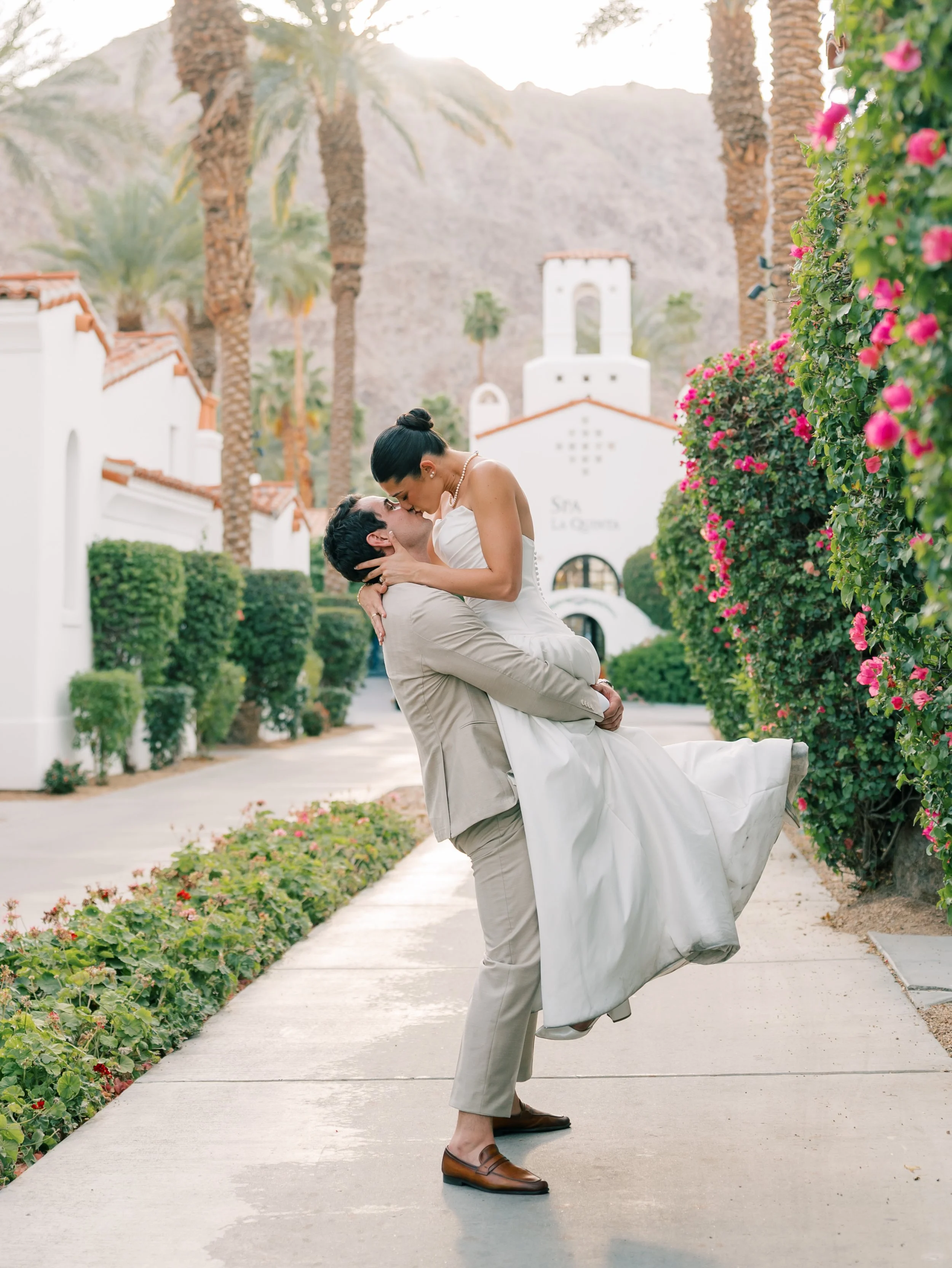 A man lifting a woman in a white dress and pearl necklace in an outdoor setting with palm trees, flowering bushes, white buildings, and a mountain in the background.