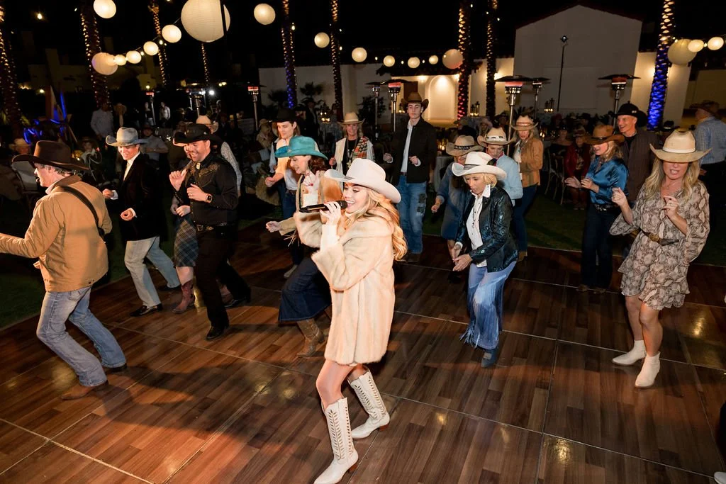 People dancing at a country-themed event with Western attire, cowboy hats, and boots, decorated with hanging paper lanterns and string lights.