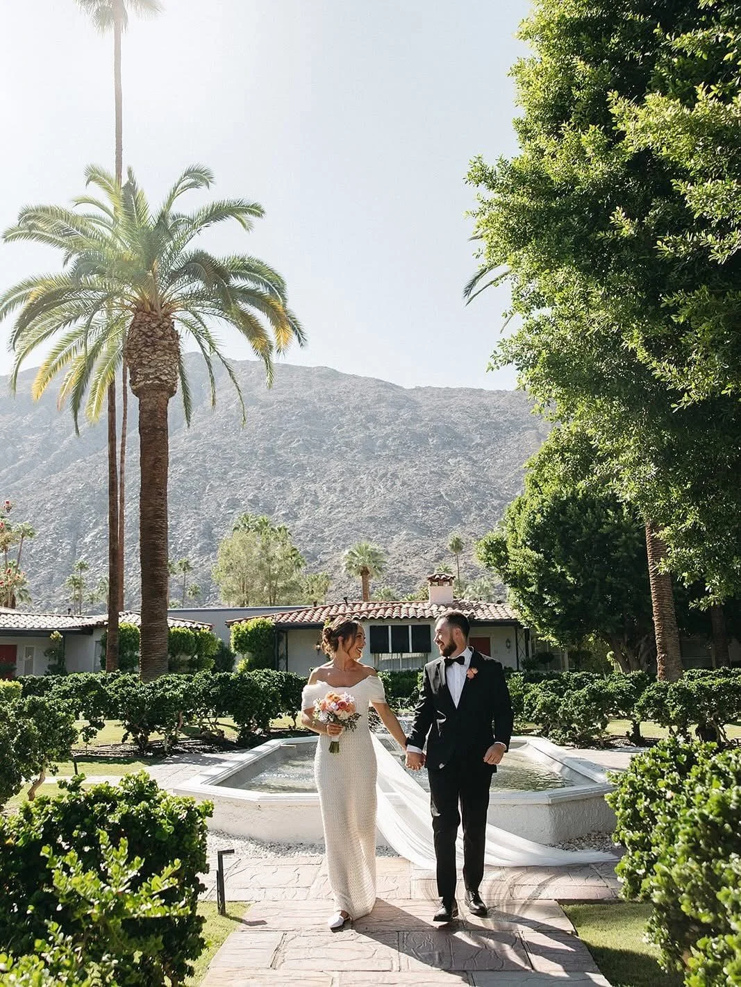 A bride in a white wedding dress holding a bouquet and a groom in a black tuxedo walking hand in hand outdoors in a garden with palm trees, bushes, and a mountain in the background.