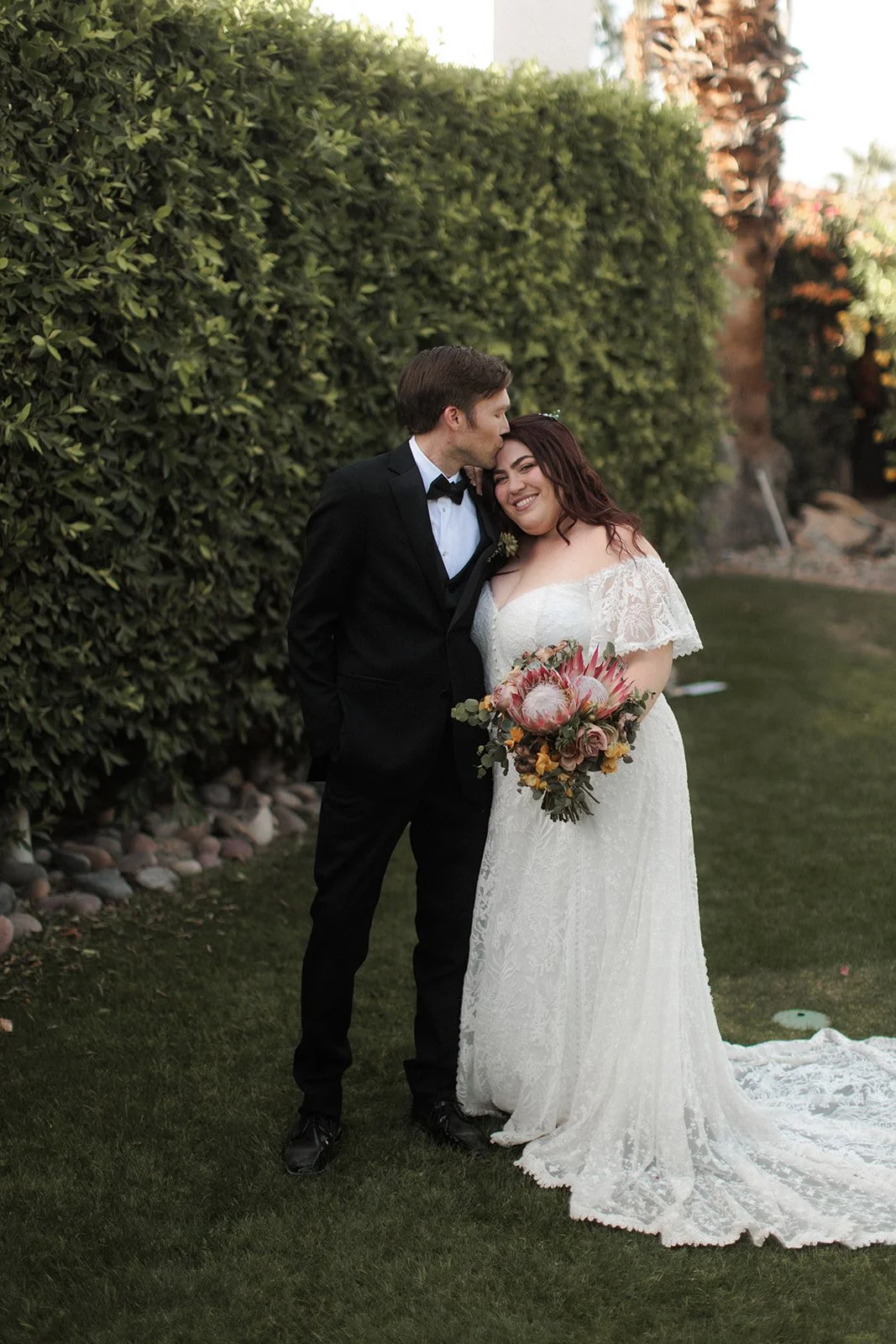 A newlywed couple standing outdoors, the groom in a black tuxedo kissing the bride on her forehead, the bride in a white lace wedding dress holding a bouquet of flowers, both smiling.