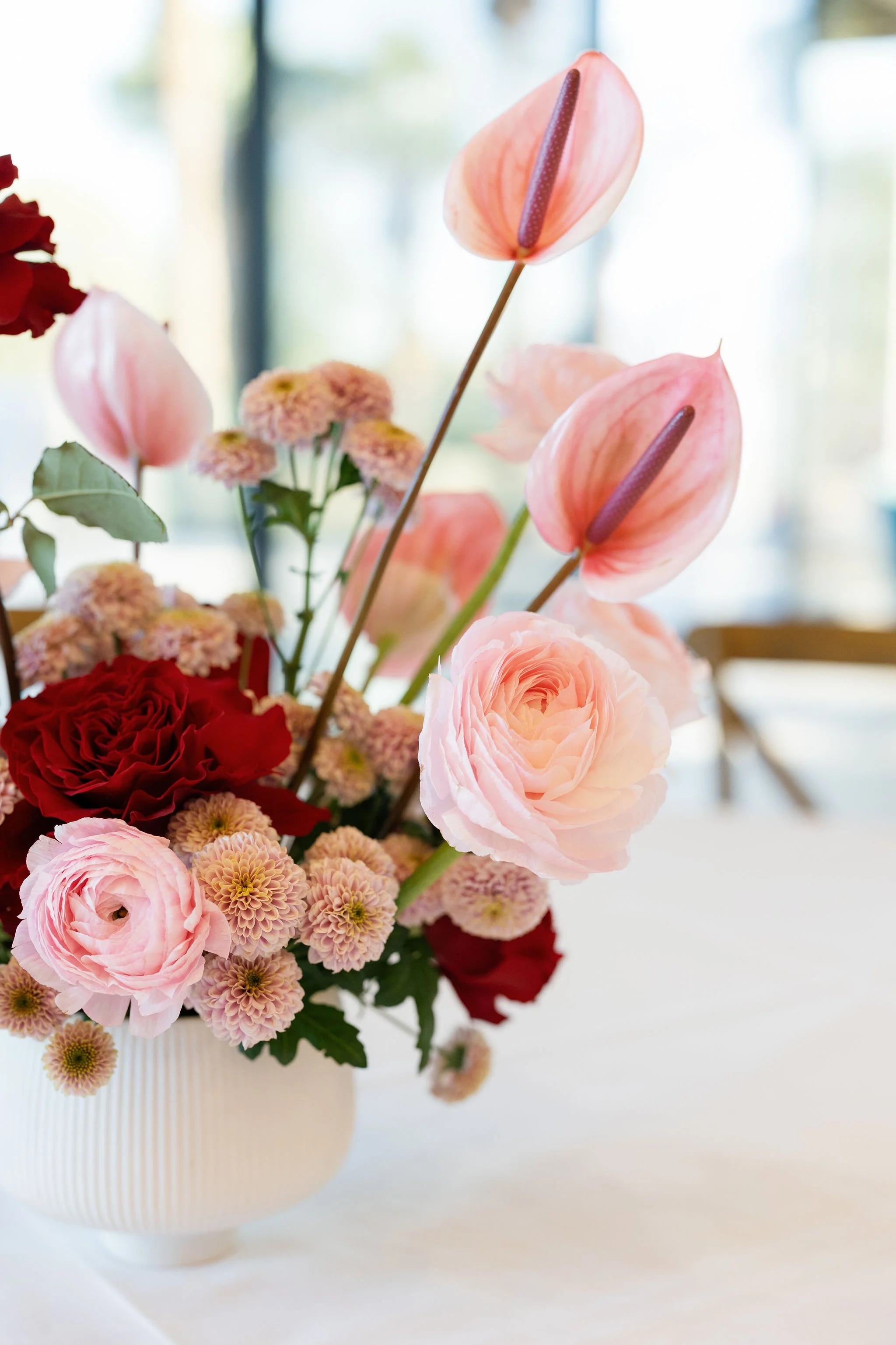 A white ceramic vase with vertical ridges holding a bouquet of pink, peach, and red flowers, including roses and anthuriums, on a white surface with a blurred background of windows and outside scenery.