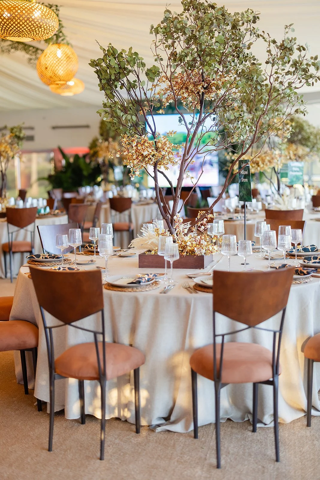 Elegant indoor banquet setup with a round table decorated with a large floral centerpiece featuring greenery and yellow flowers, set with wine glasses, plates, cutlery, and napkins, surrounded by wooden chairs with peach cushions, and warm lighting w