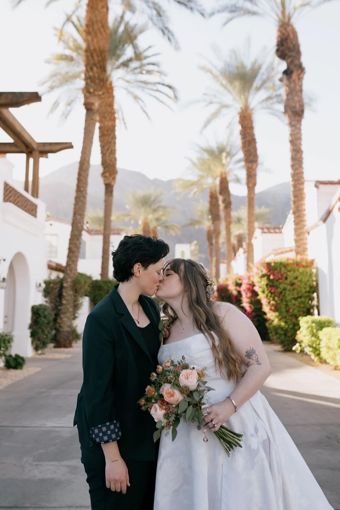 Two brides share a kiss. The bride in the white wedding dress holds a beautiful bouquet of flowers. The background shows a stunning desert resort landscape with palm trees and mountains.