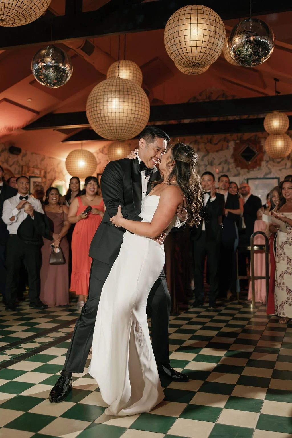 A bride and groom dance under lanterns and disco balls in a beautiful pink room on a green and white checkered dance floor.