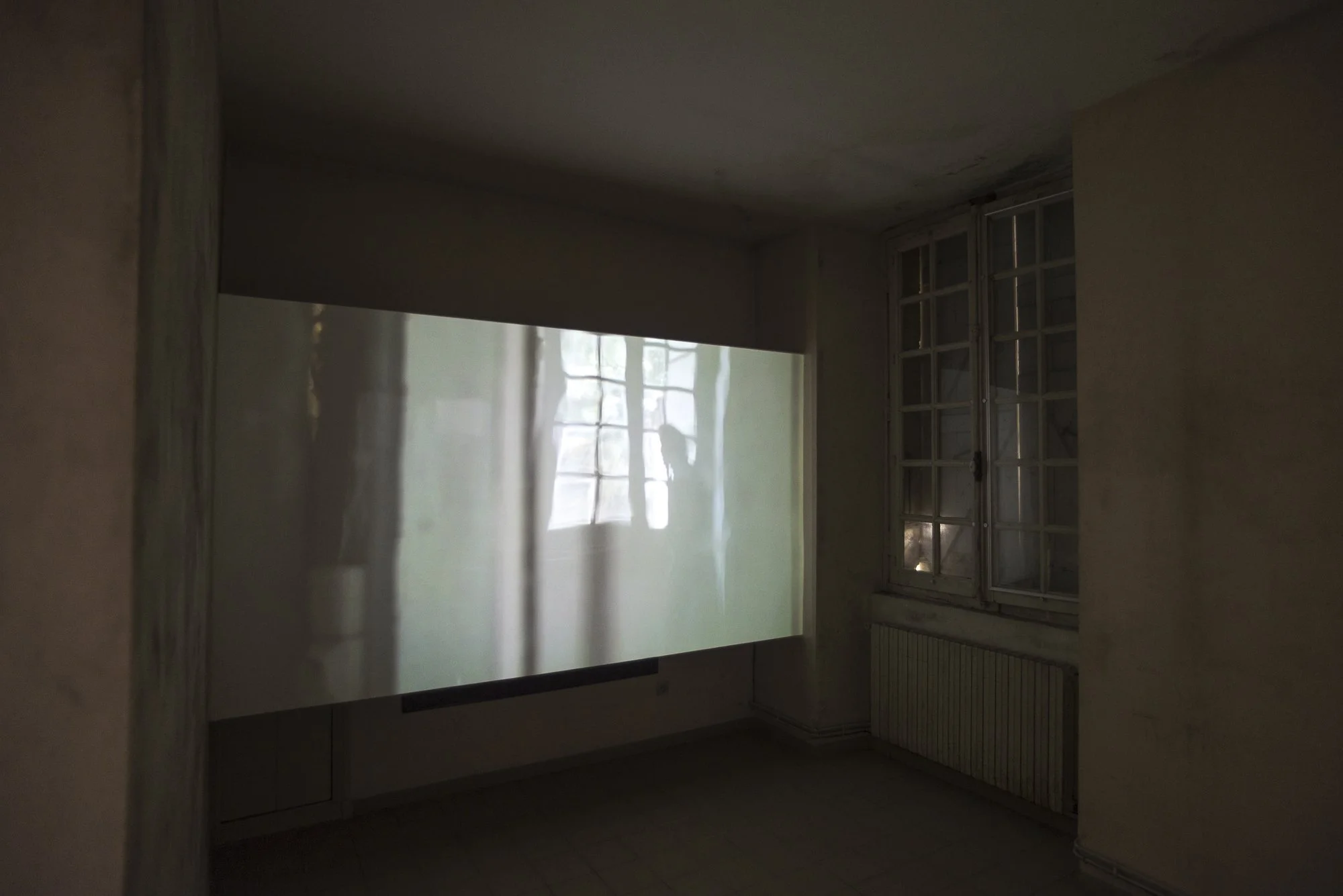 An empty room with a window on the right and a projected screen or wall on the left, with faint reflections of window panes and trees visible.