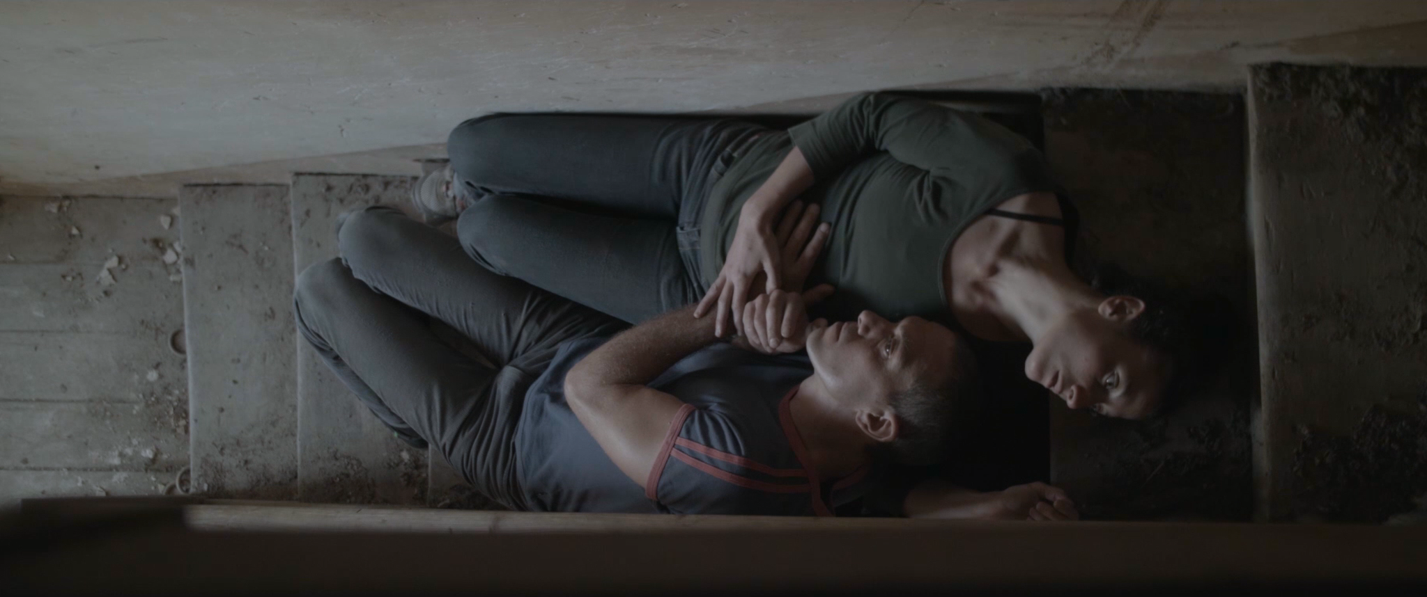 Two people, a man and a woman, lying on the ground in a narrow space, holding hands and looking into each other's eyes. The woman is wearing a green shirt and dark pants, the man is wearing a gray T-shirt and dark pants.