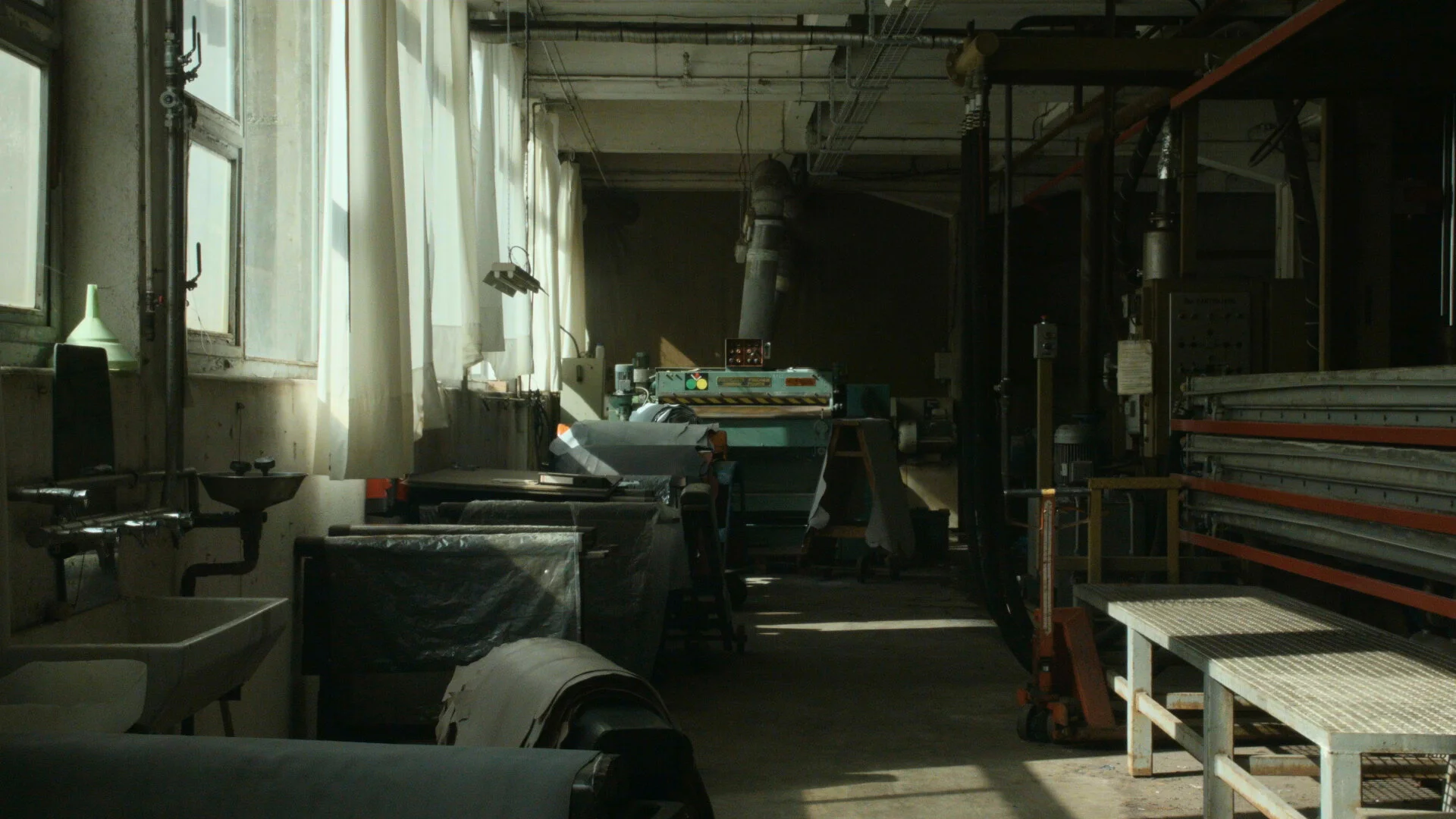 An industrial workshop with machinery, workbenches, and tools, illuminated by natural sunlight through large windows with white curtains.