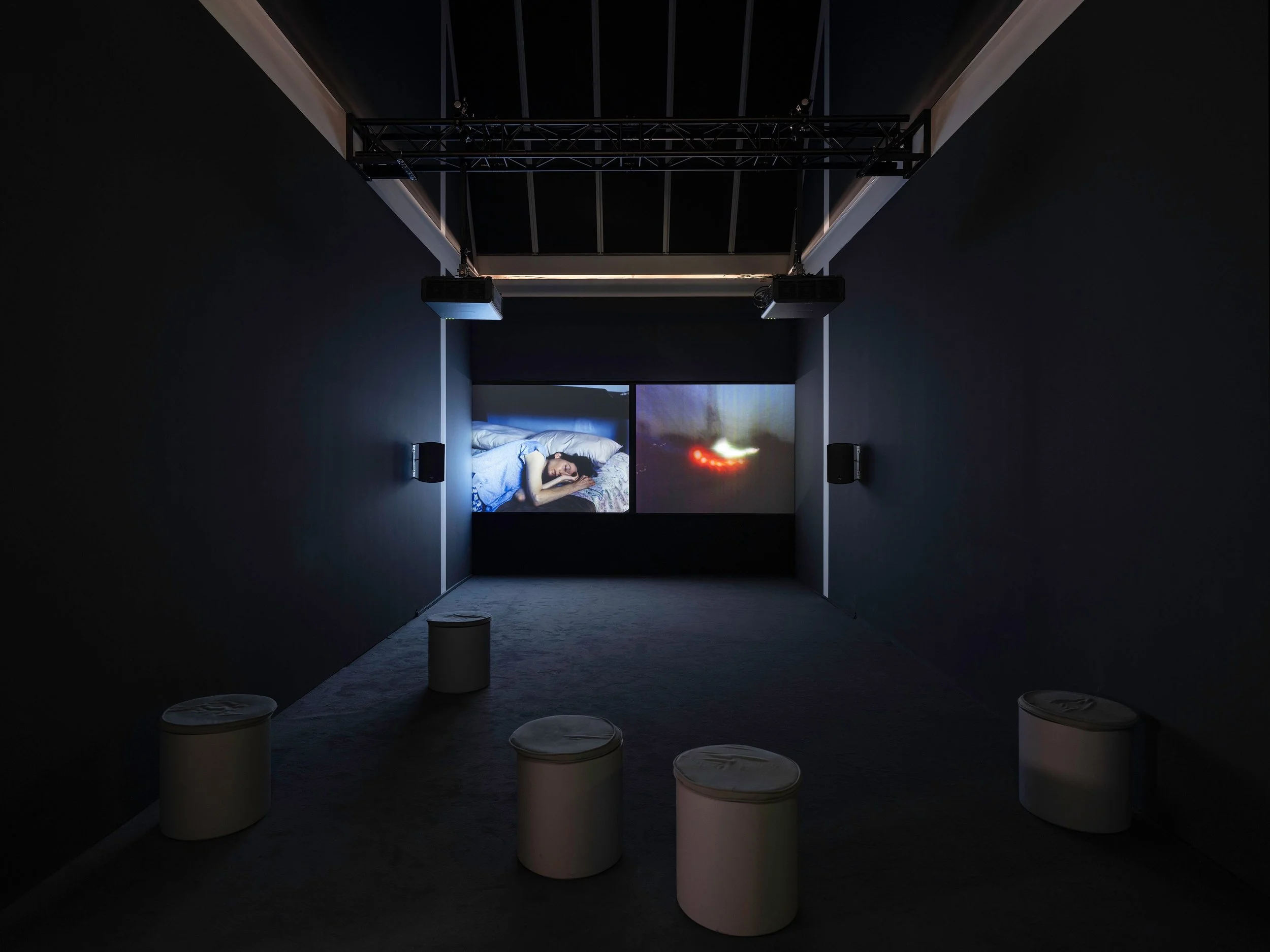 A dark room with a woman sleeping on a bed projected on a screen, with another screen showing a spaceship. There are four white stools in the room.