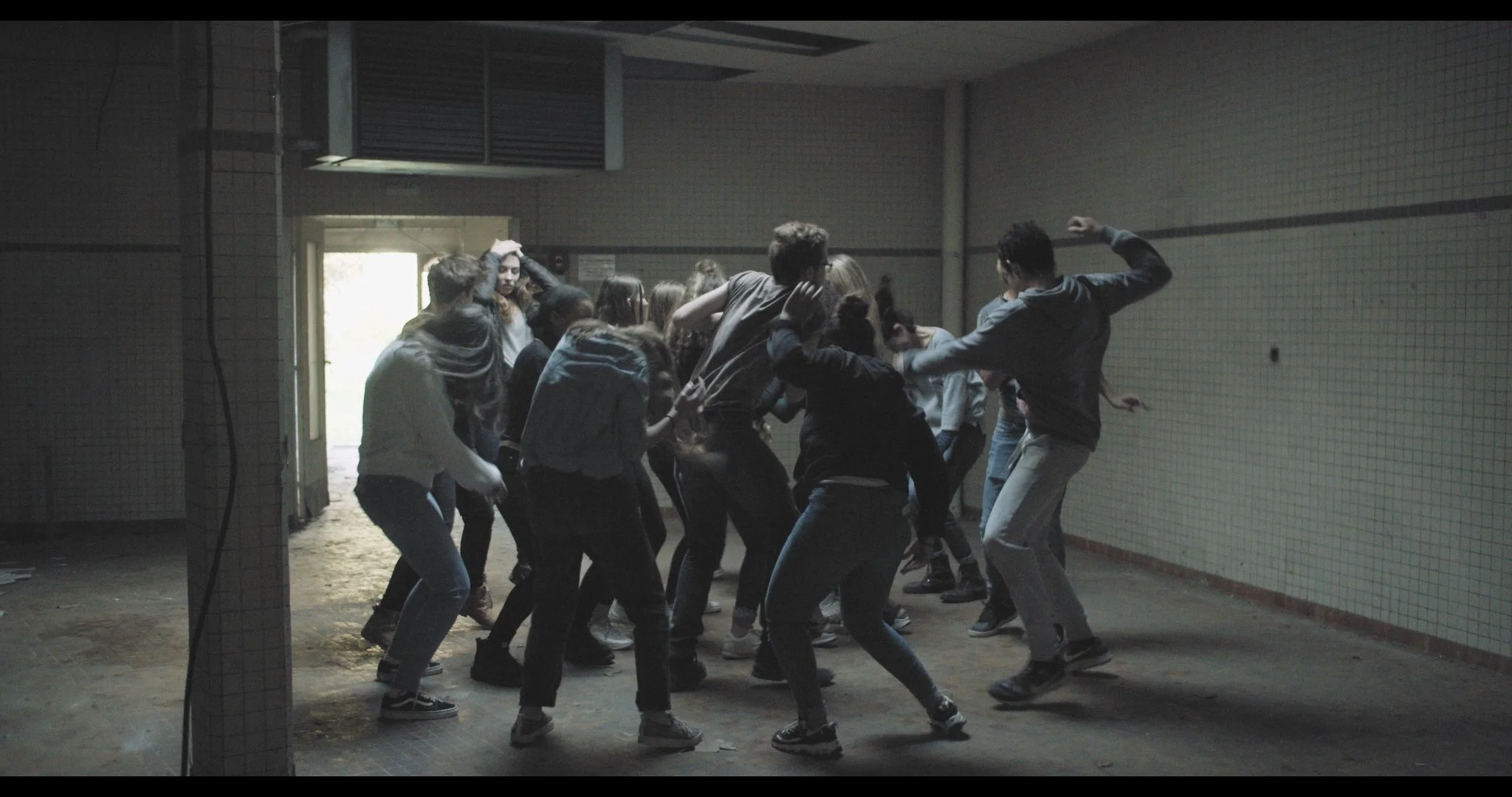 A group of people are dancing or fighting in an abandoned, dimly lit room with tiled walls and a concrete floor, with a light source coming from the door on the left.