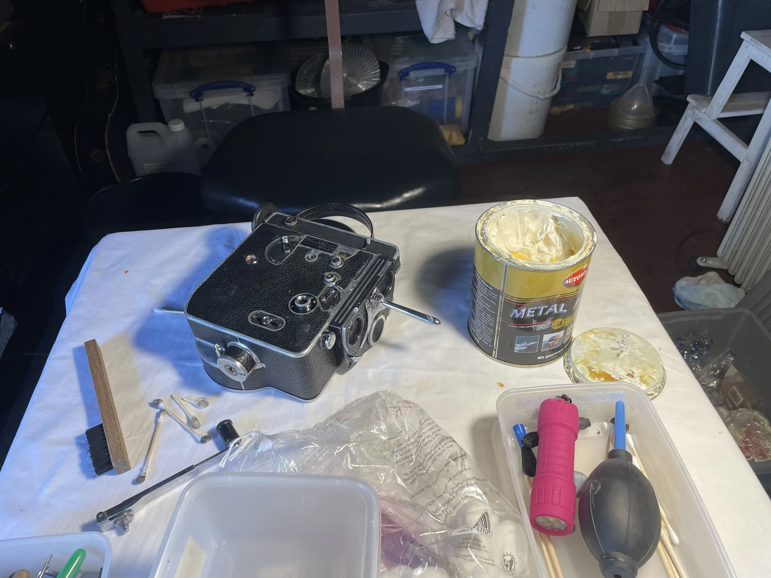 A workbench with a vintage film camera, a can of metal primer with yellowish paint, a pink flashlight, a computer mouse, some small tools, a wooden stick, and miscellaneous items in a garage workspace.