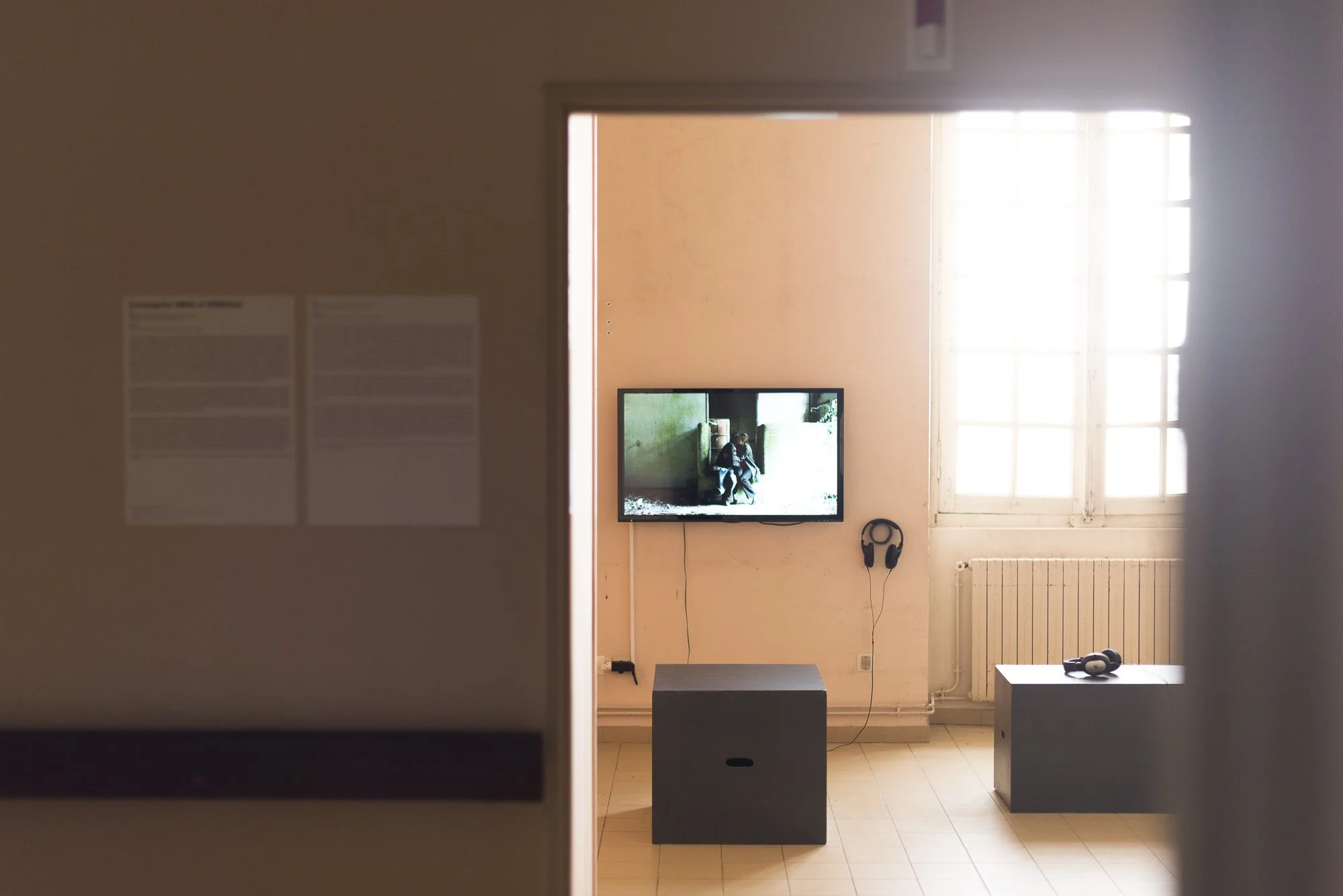 A room with a wall-mounted television, headphones hanging on a hook, a small table with headphones on it, a large window letting in natural light, and a radiator below the window.