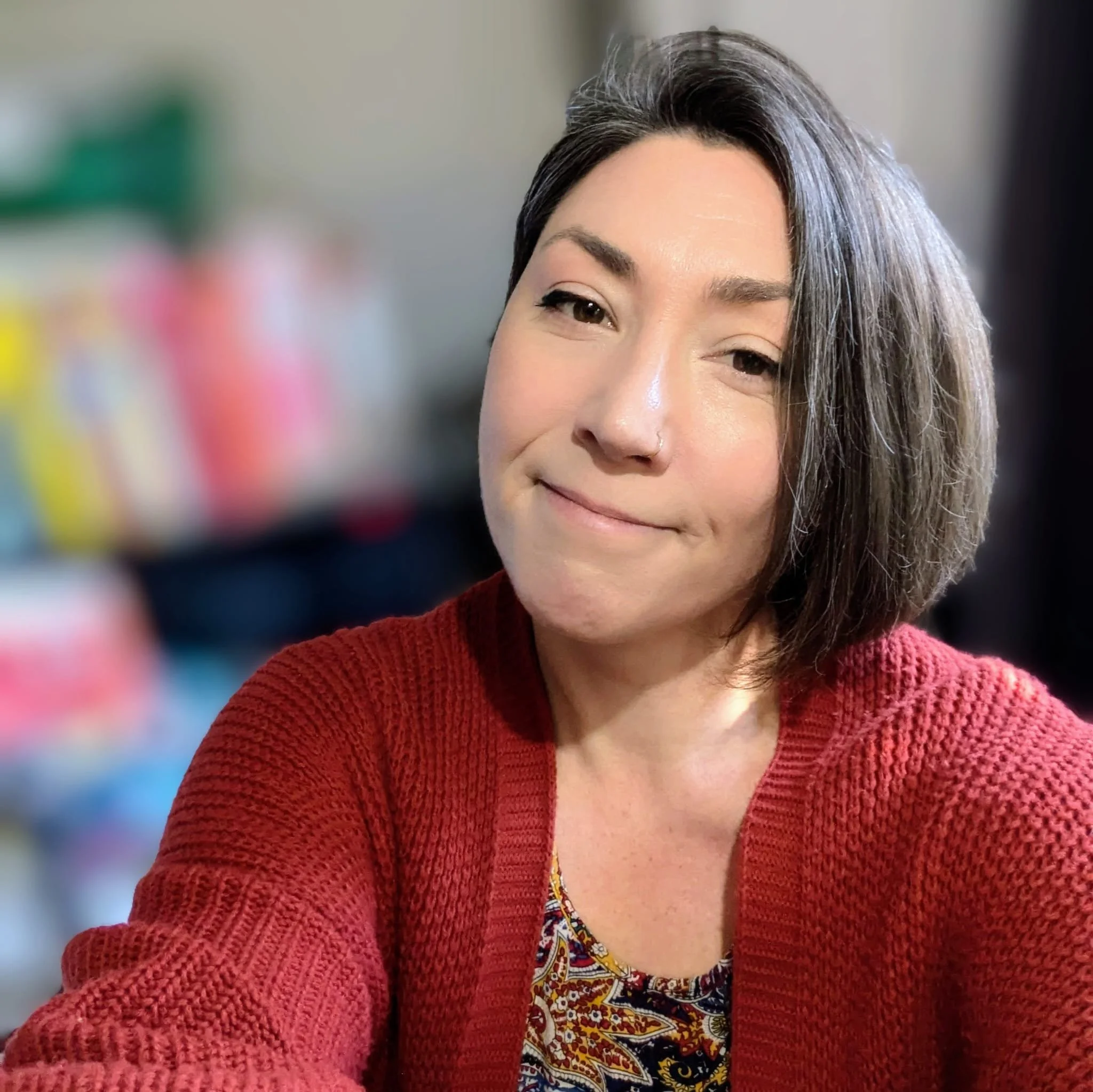 A woman with short brown hair wearing a red sweater and patterned shirt, smiling slightly, indoors with blurred colorful objects in the background.