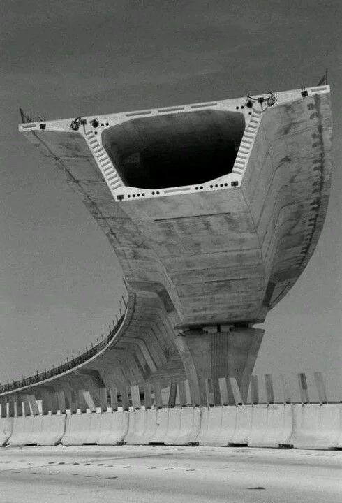 A large construction site featuring a massive, partially constructed concrete bridge with a tunnel under the road, viewed from below with barriers along the roadway.