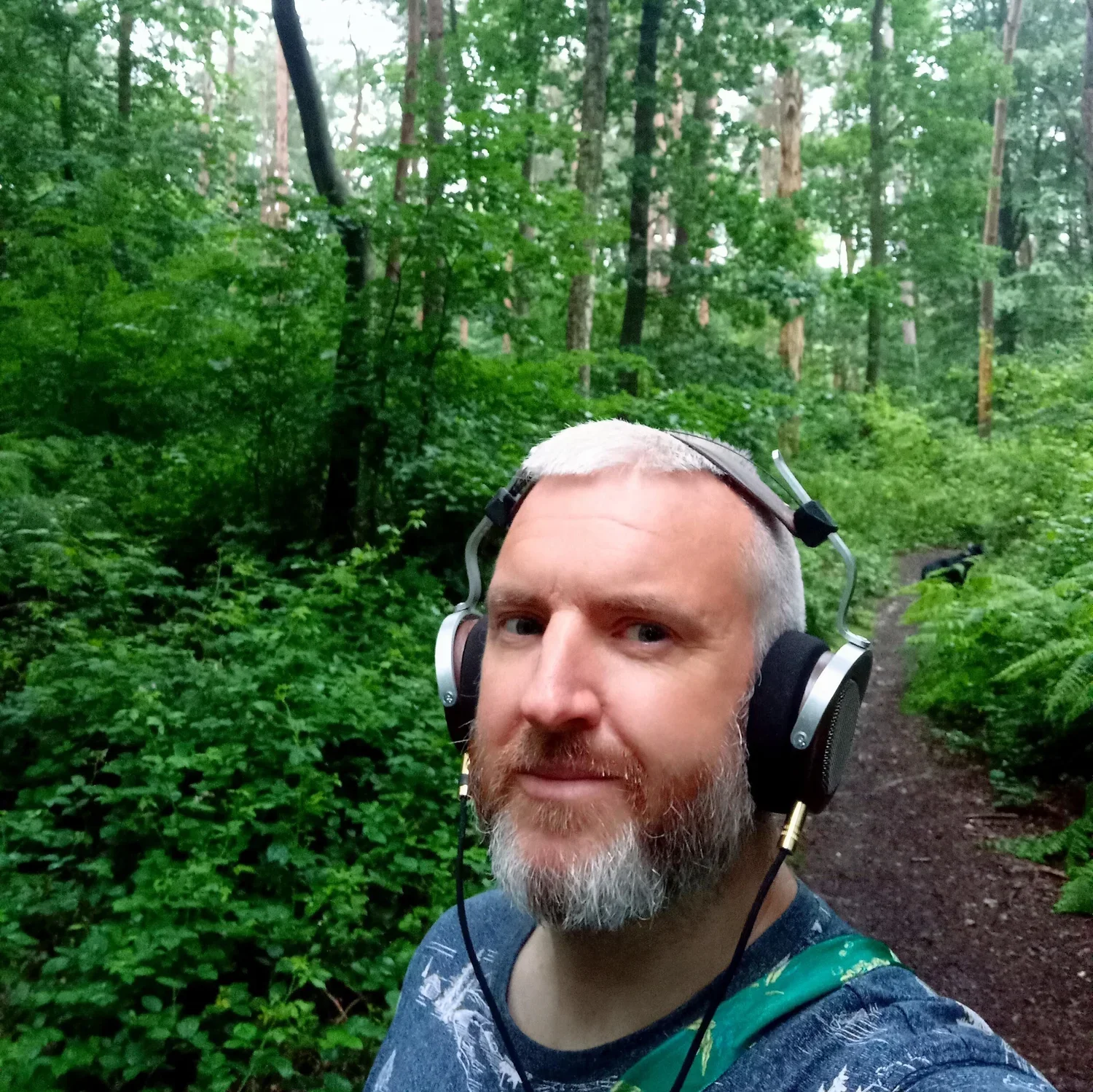 A man with short gray hair and a beard, wearing headphones, taking a selfie in a lush green forest with a dirt trail.