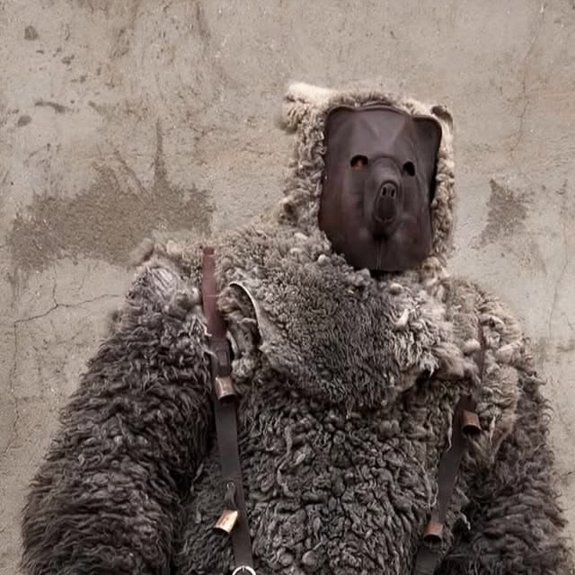 A person wearing a bear costume with a furry body and a mask with a bear face, standing against a rough wall background.