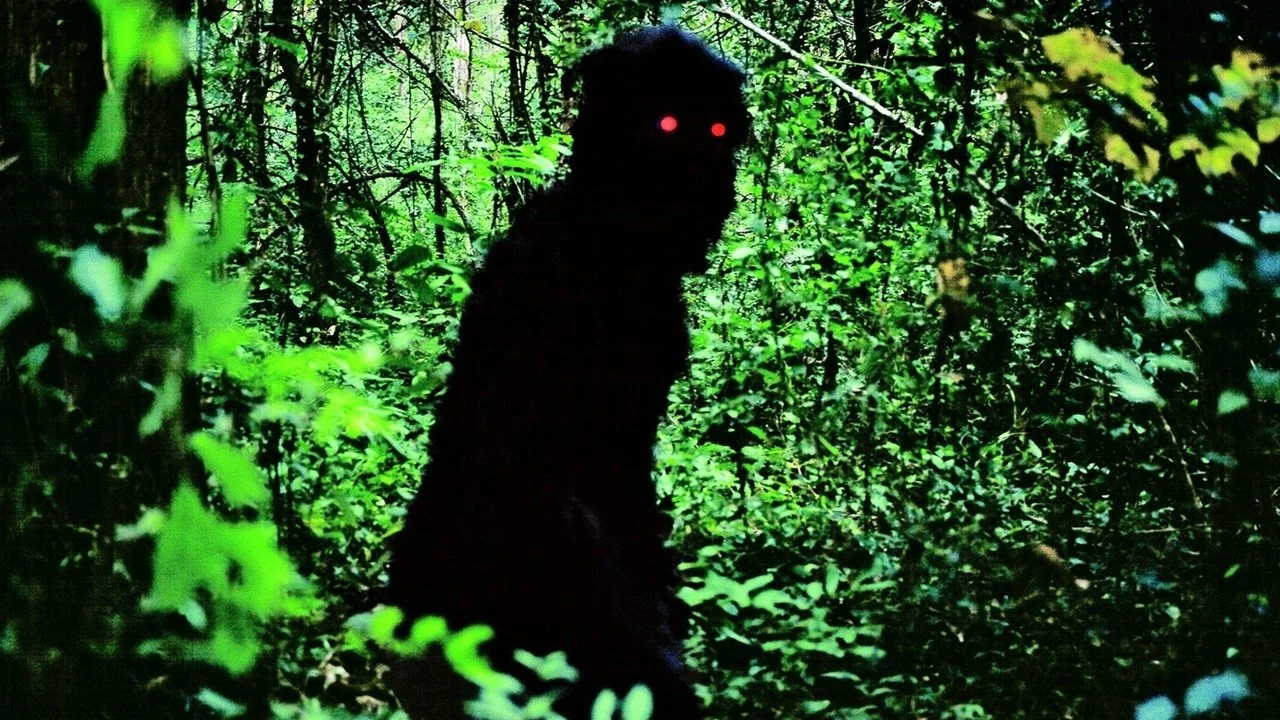 A dark figure with glowing red eyes in a dense, green forest at night.