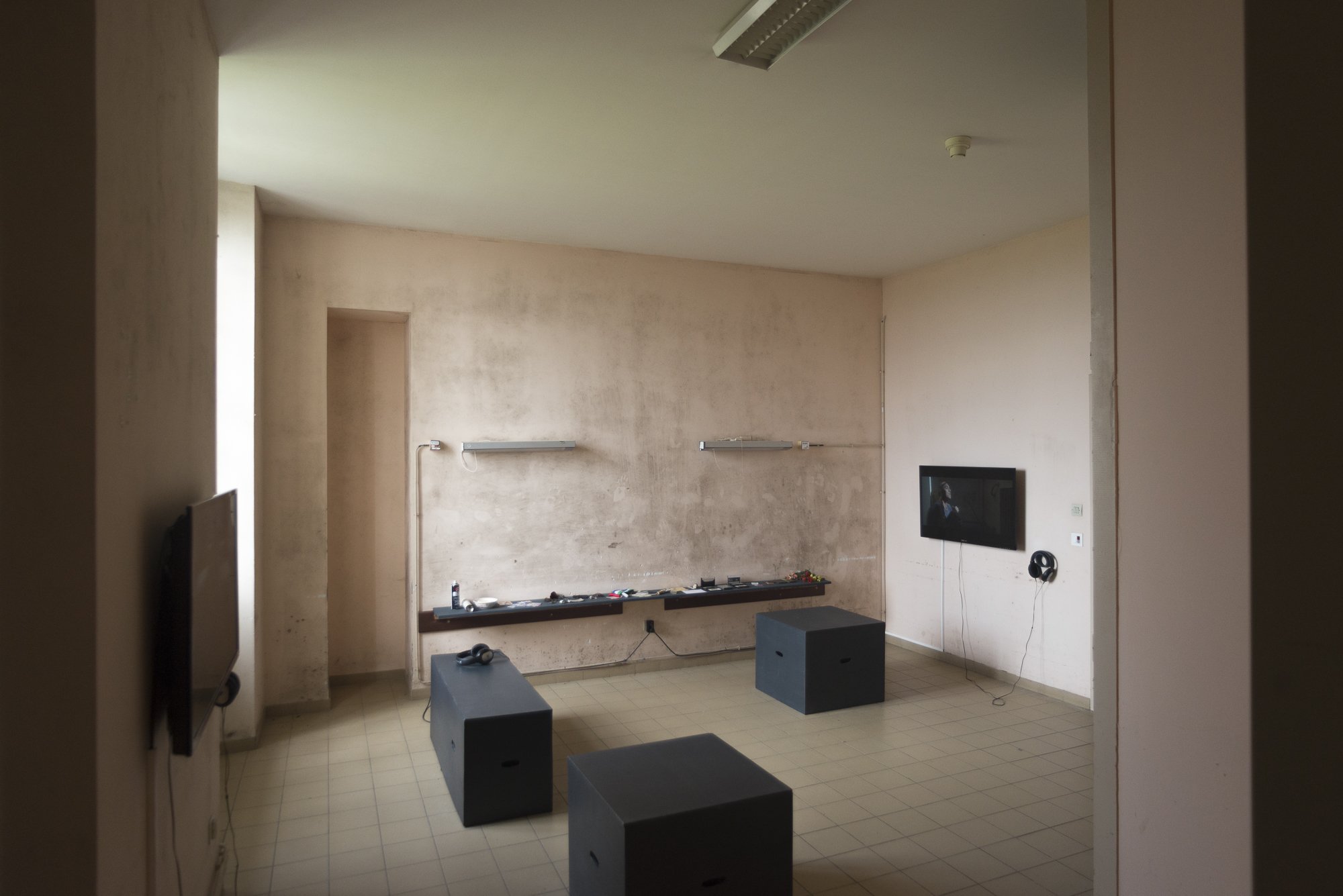 Empty living room with beige walls, mounted TV, and black furniture including three rectangular seats and a low shelf with miscellaneous items.