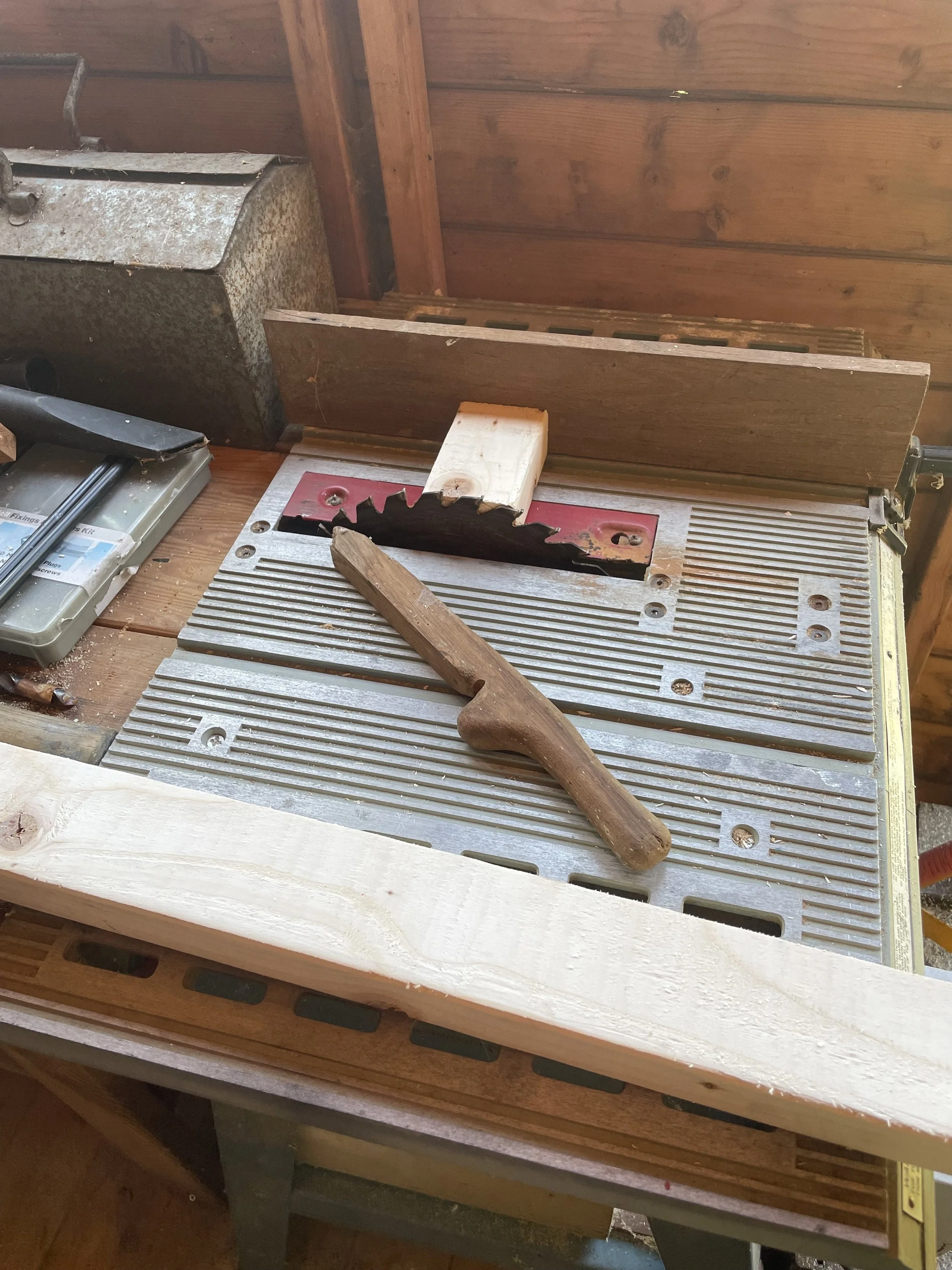 Workshop scene with a saw on a table, a hammer, and a wooden piece. The saw has a jagged blade, and the hammer is placed on a workbench.